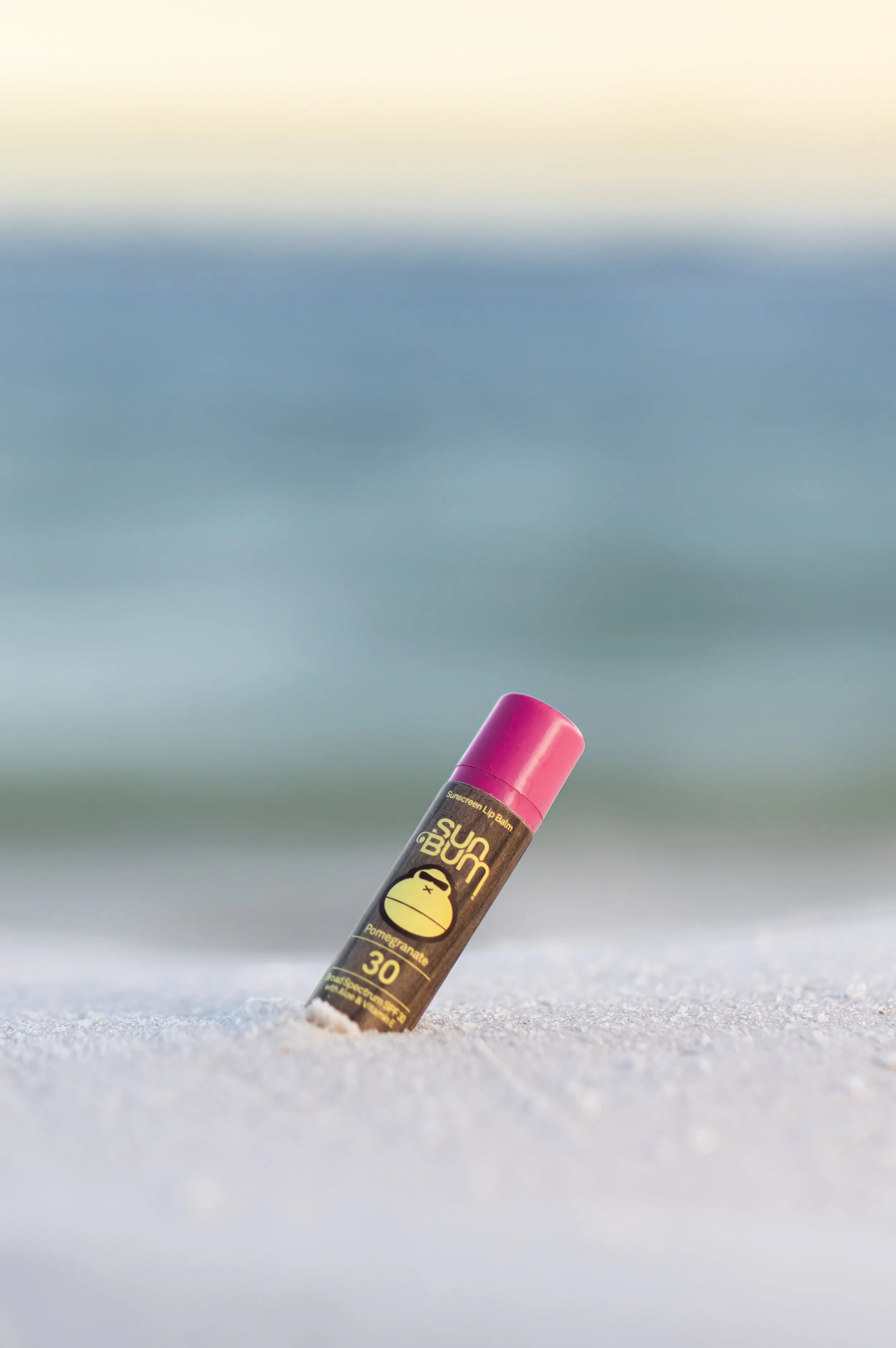 Lip balm tube labeled 'Sun Bum' with SPF 30, partially buried in the sand on a beach, with ocean and sky in the blurred background.