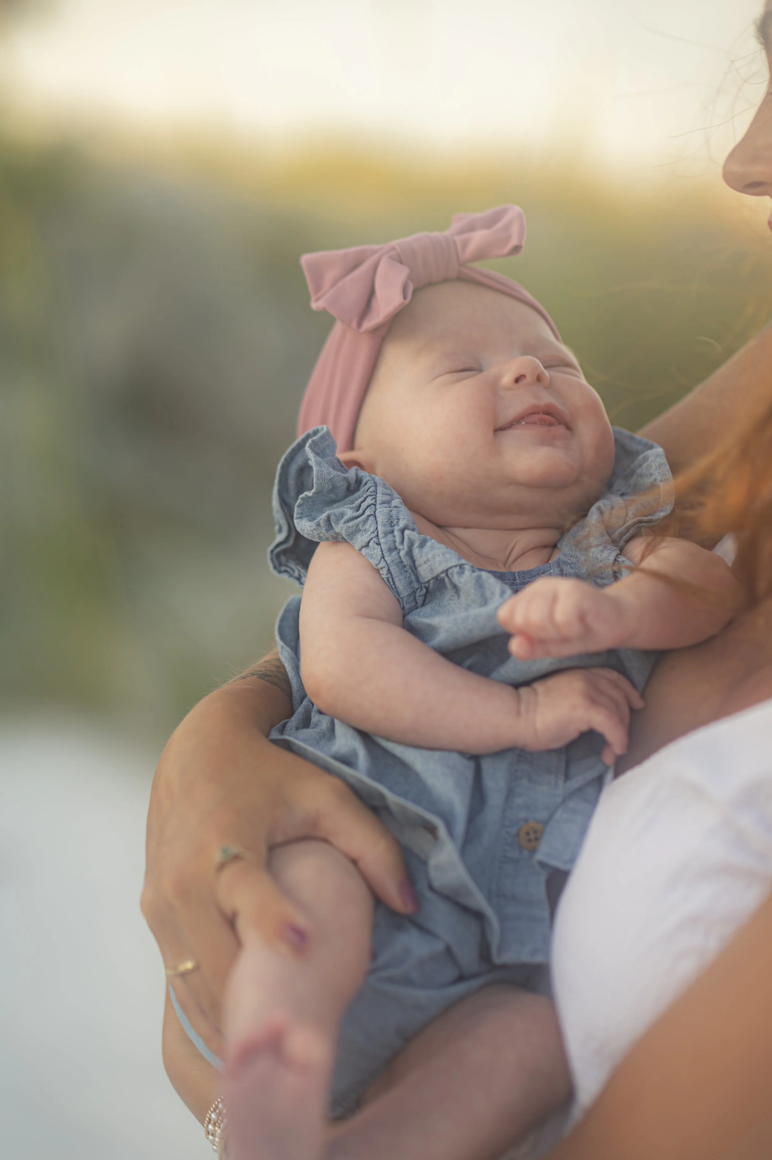 A smiling baby girl being held by an adult woman, wearing a pink headband with a bow and a denim dress, outdoors in warm lighting.