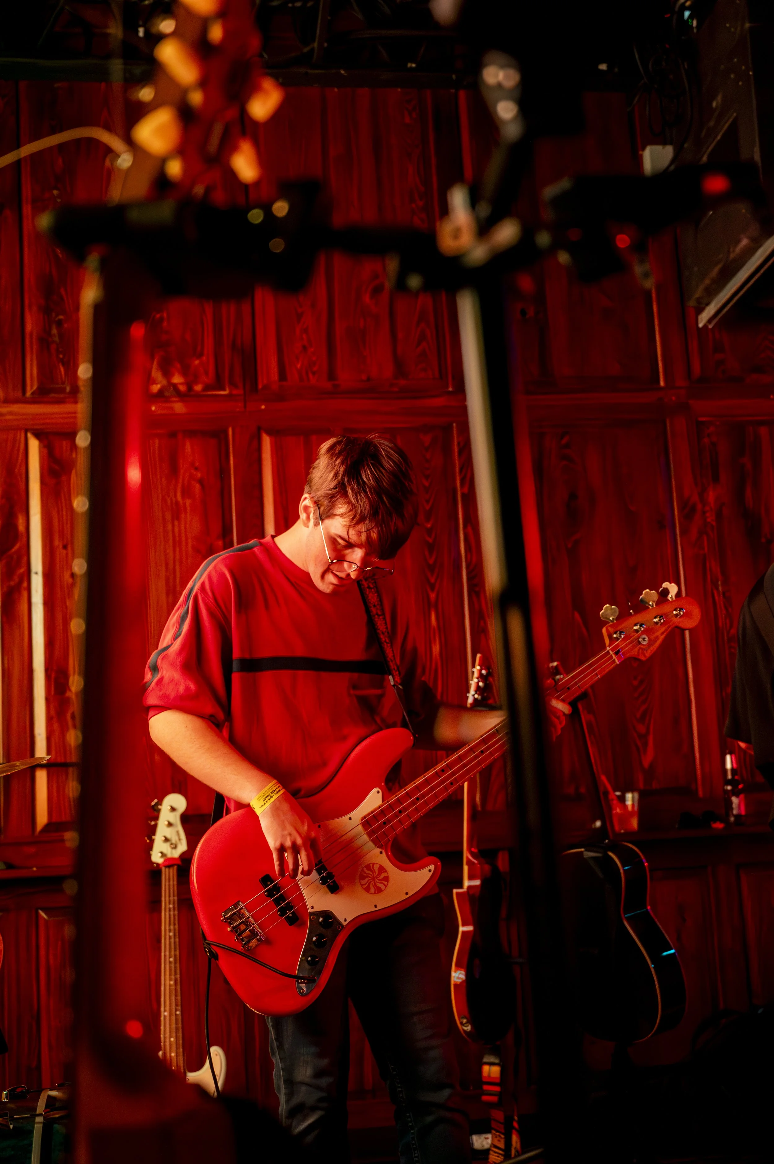 A young man playing a red electric bass guitar during a live performance in a wood-paneled room.