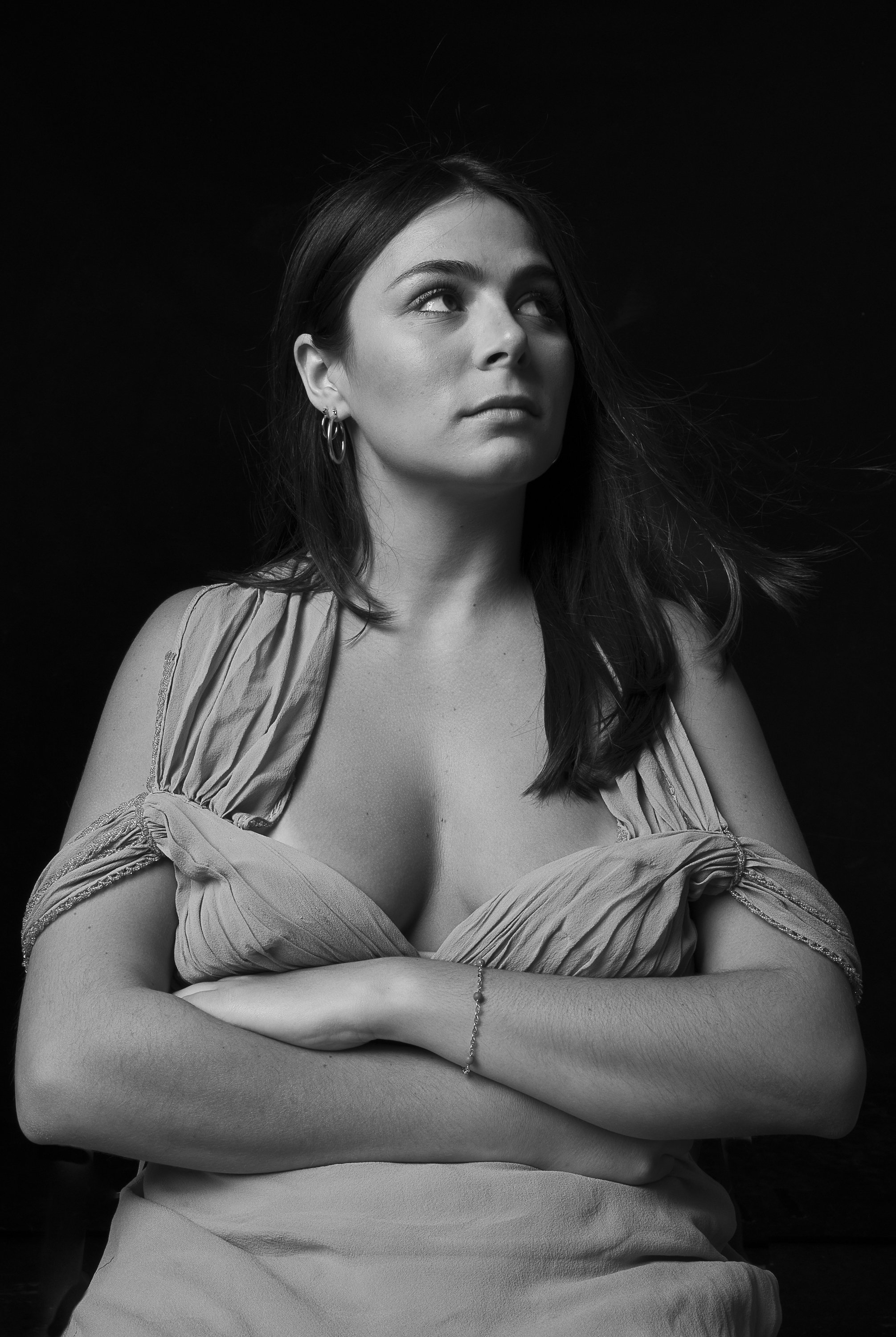 A young woman with dark hair, wearing earrings and a dress with puffed sleeves, looking upwards and crossing her arms, in black and white studio portrait.