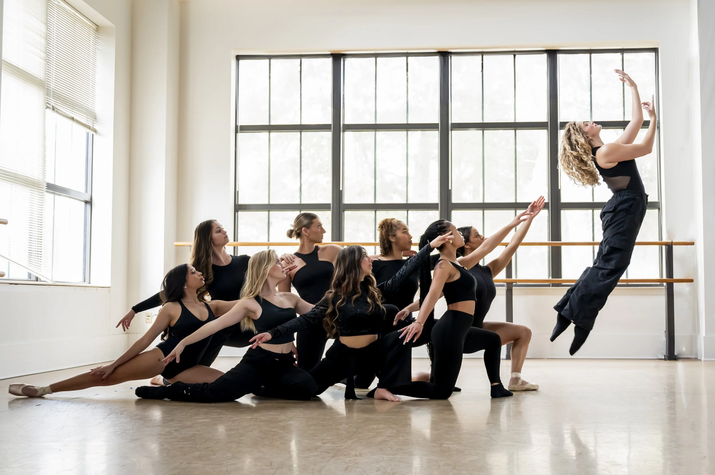 A group of nine female ballet dancers in black dancewear practicing in a dance studio with large windows and a wooden barre.