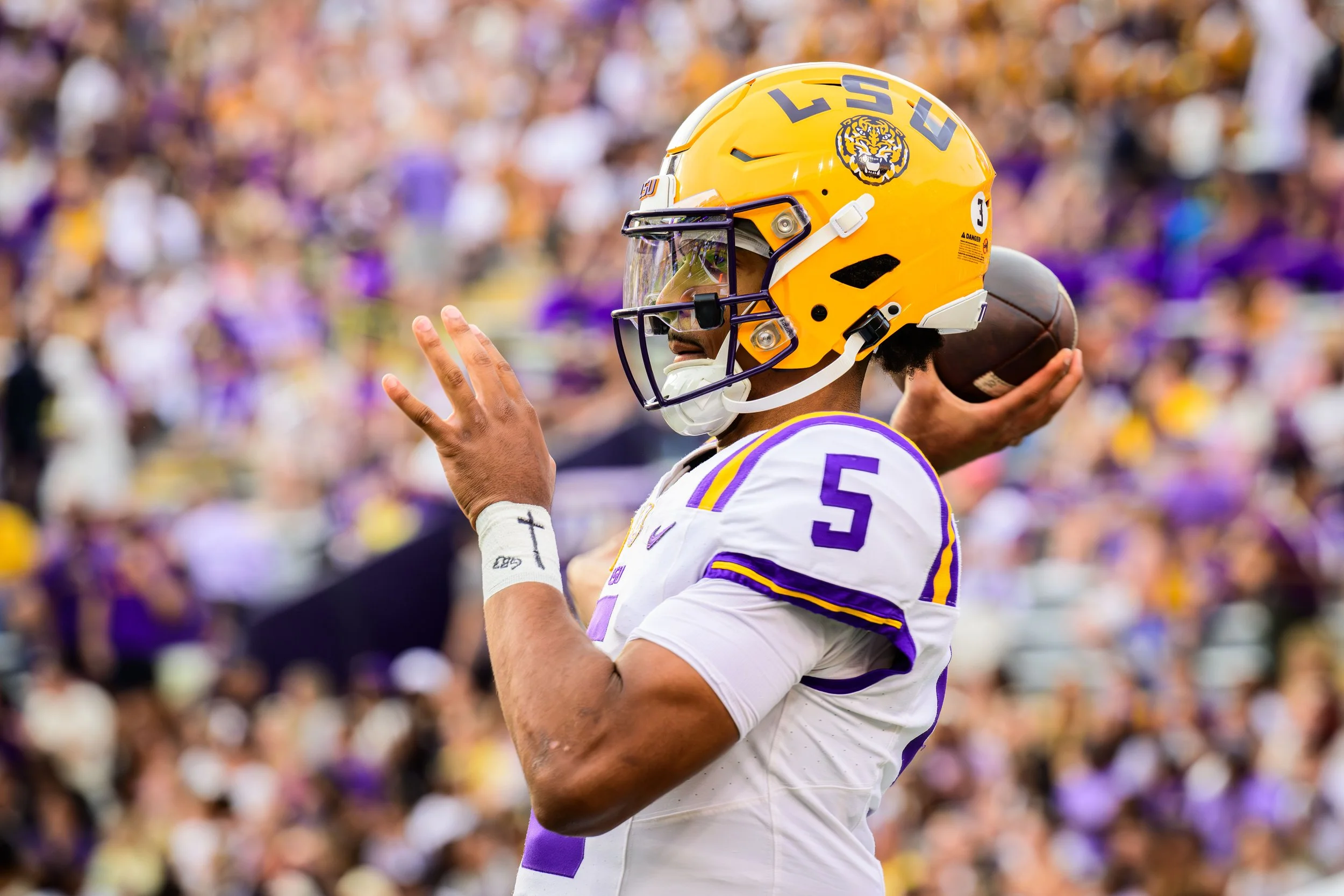 An LSU football player in a white uniform with purple and yellow accents, wearing a yellow helmet with a tiger logo, holding a football and preparing to throw during a game.