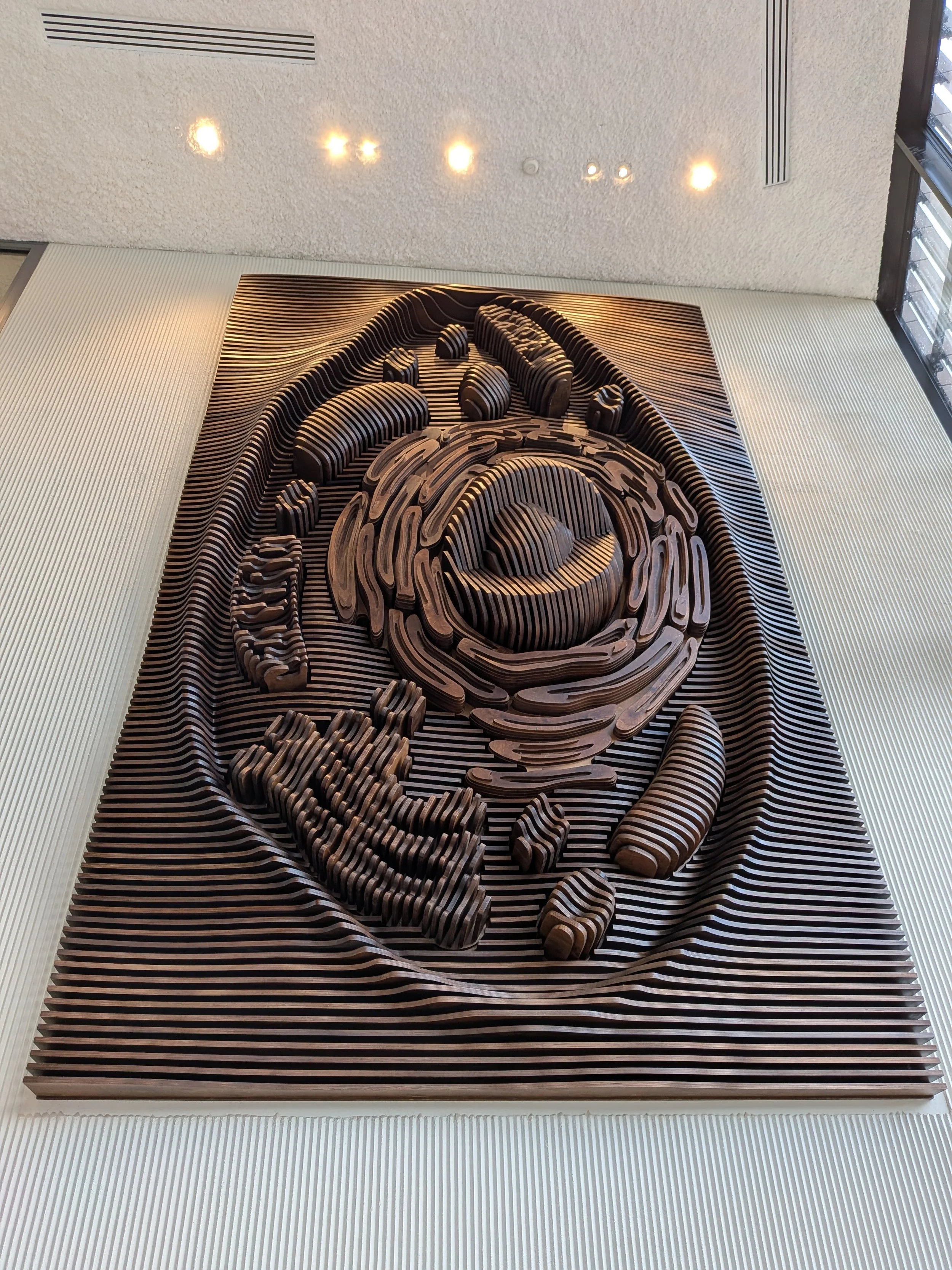 A large wooden sculptural piece featuring a spiral pattern and intricate details, mounted on a wall with vertical striped background in a modern interior space.