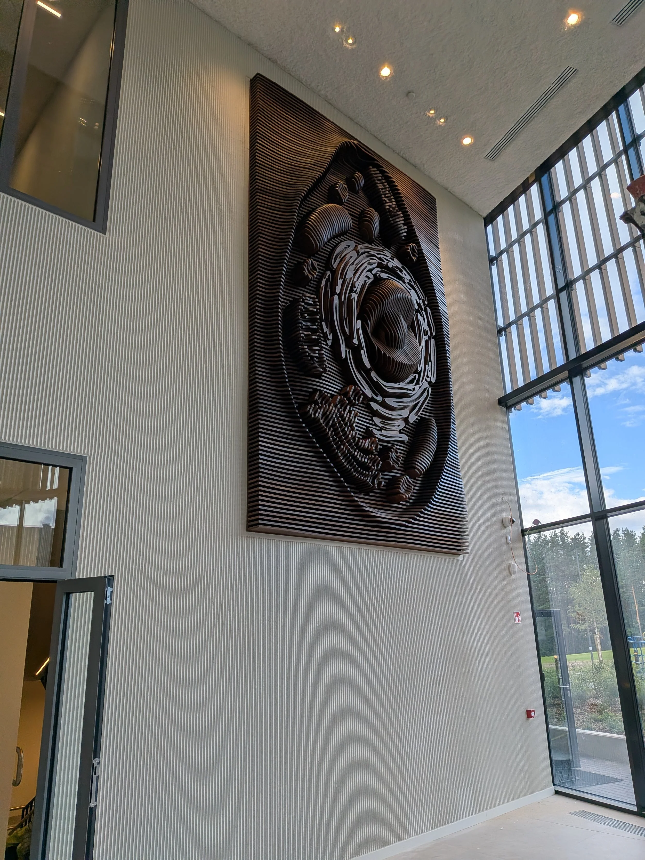 Large art installation with layered wooden panels forming a stylized face or mask on a beige wall in a modern building.
