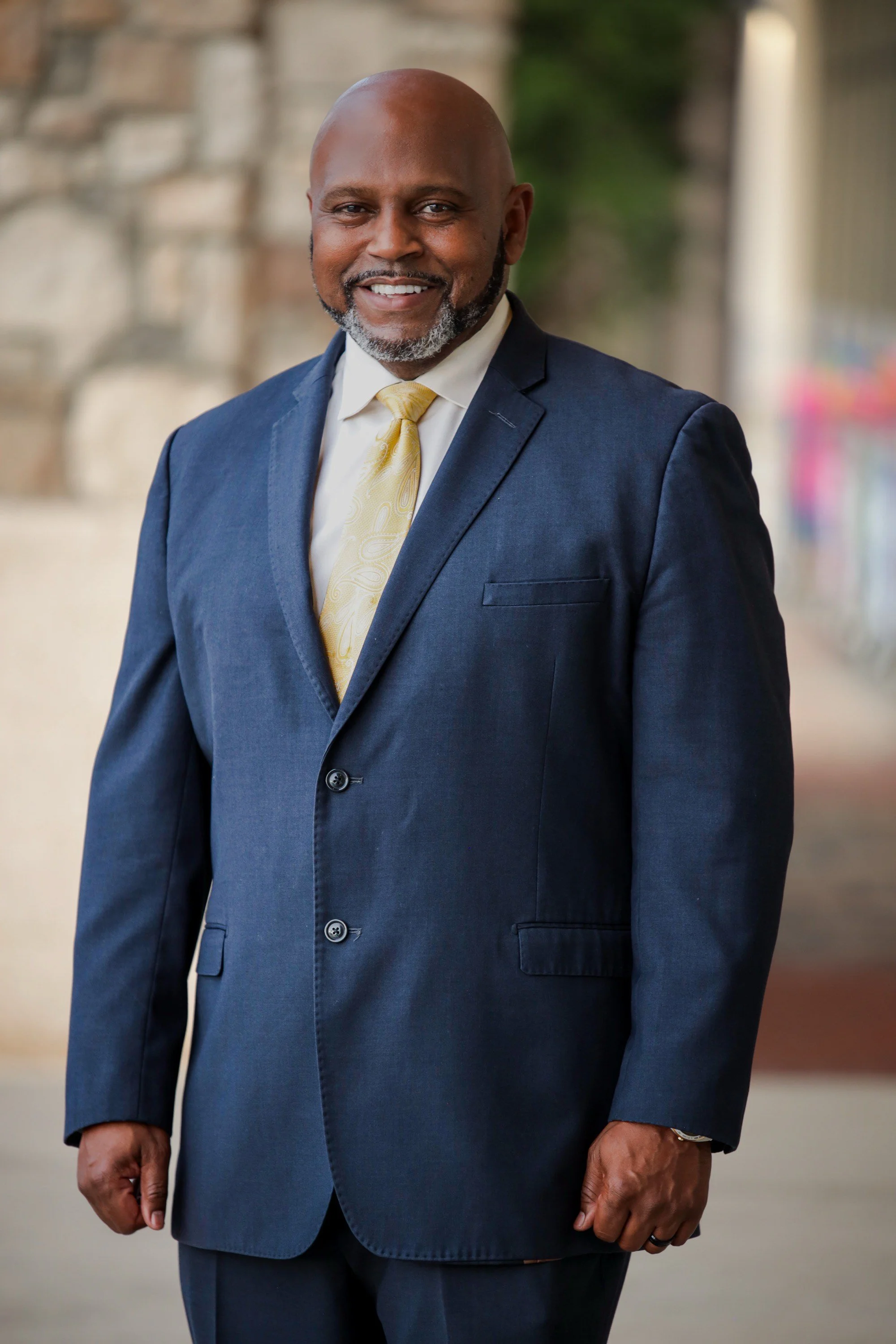 A smiling man in a navy suit, white shirt, and yellow tie standing outdoors with a blurred background.