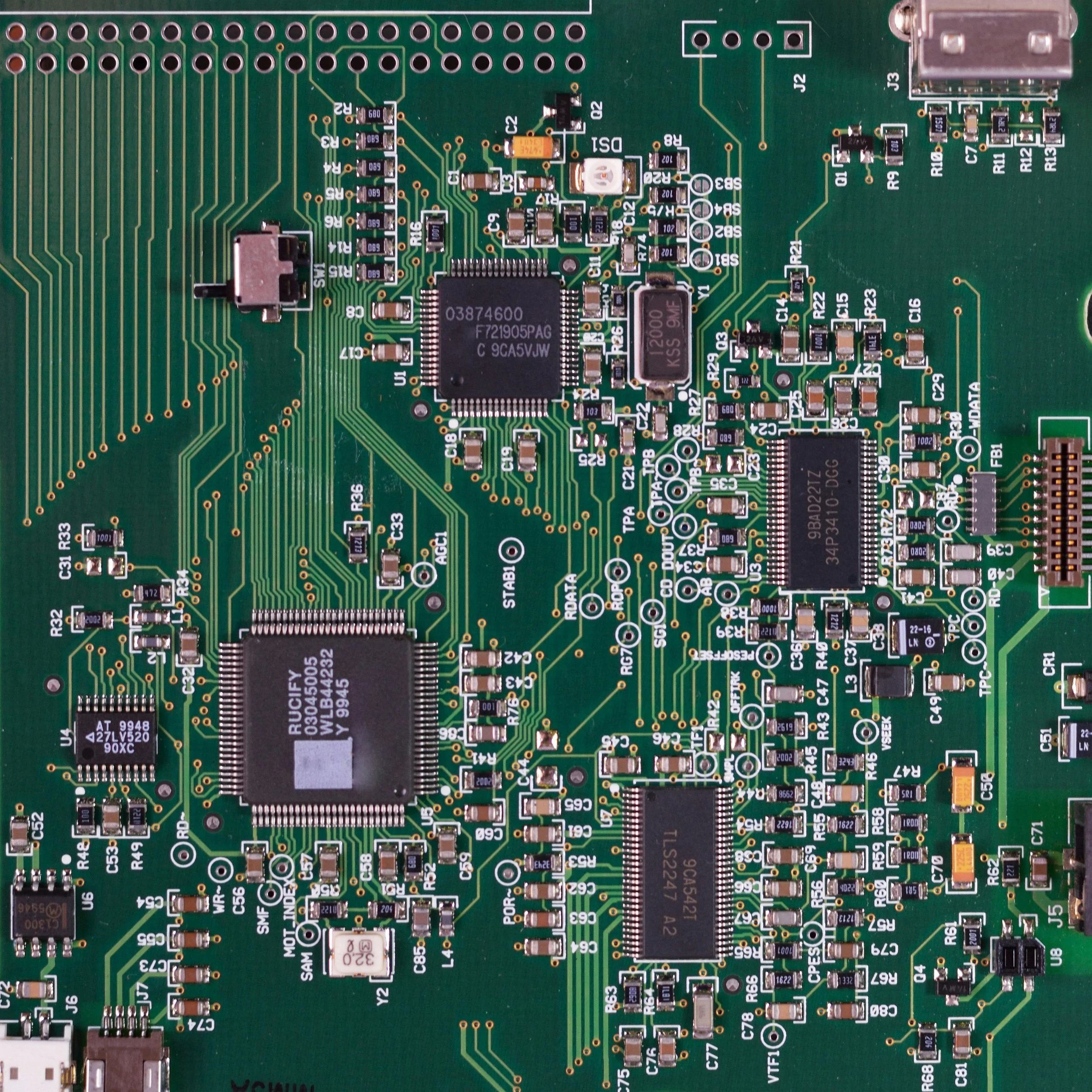 Close-up of a green printed circuit board with various electronic components including chips, resistors, capacitors, and connectors.