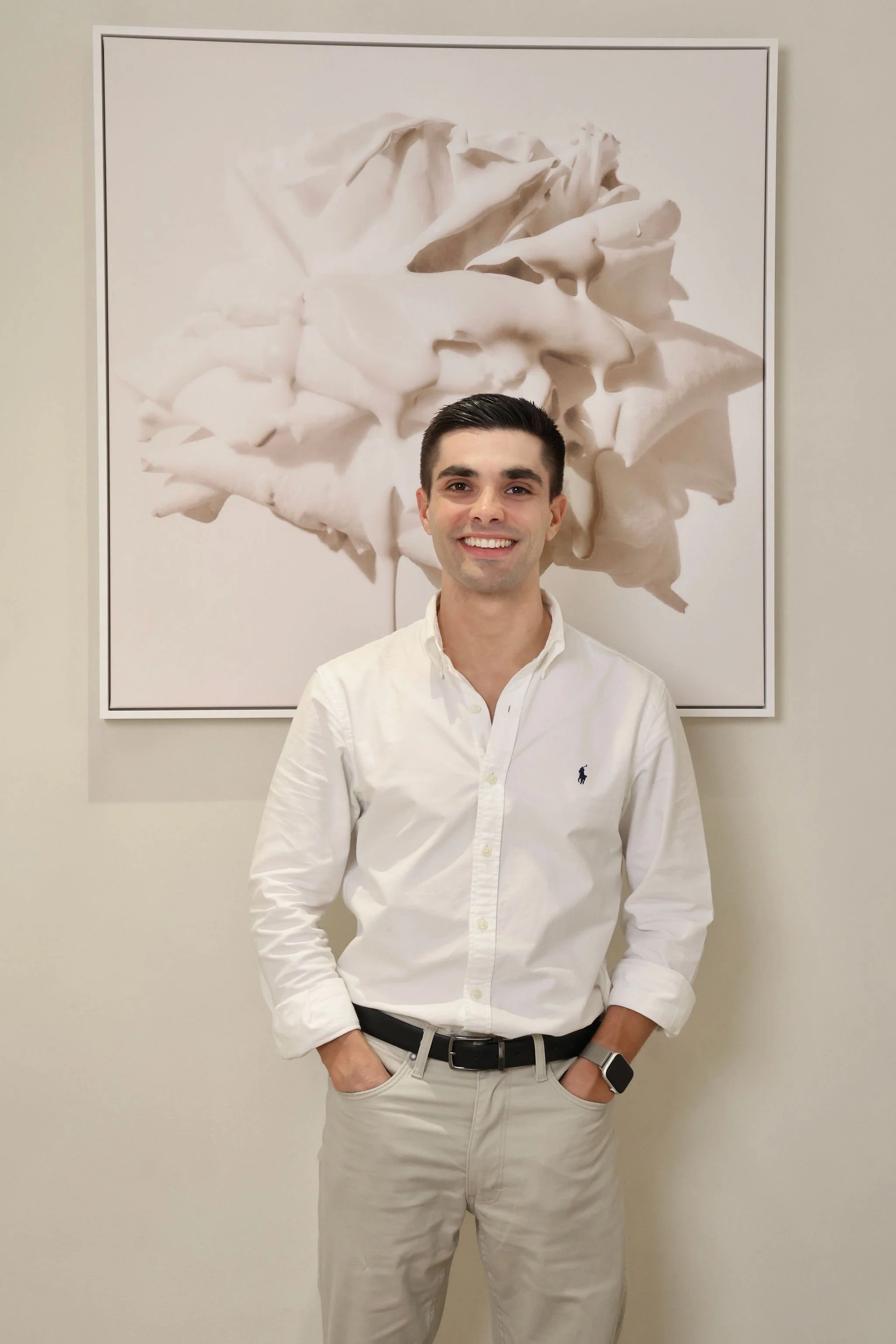 A young man dressed in a white button-up shirt and light-colored pants stands in front of a large framed abstract floral painting, smiling with his hands in his pockets.