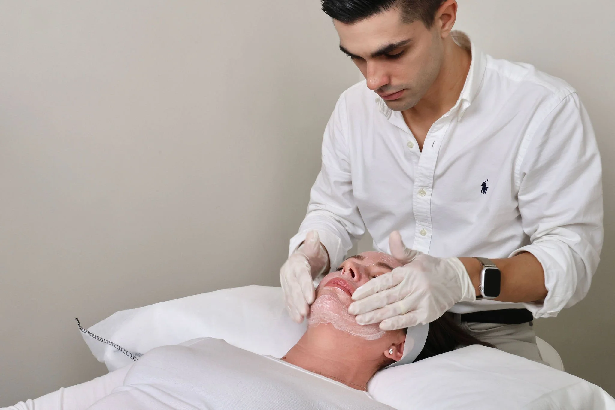 A man in a white shirt and watch is applying a facial treatment to a woman lying on a bed. The woman has a facial mask on and appears relaxed.