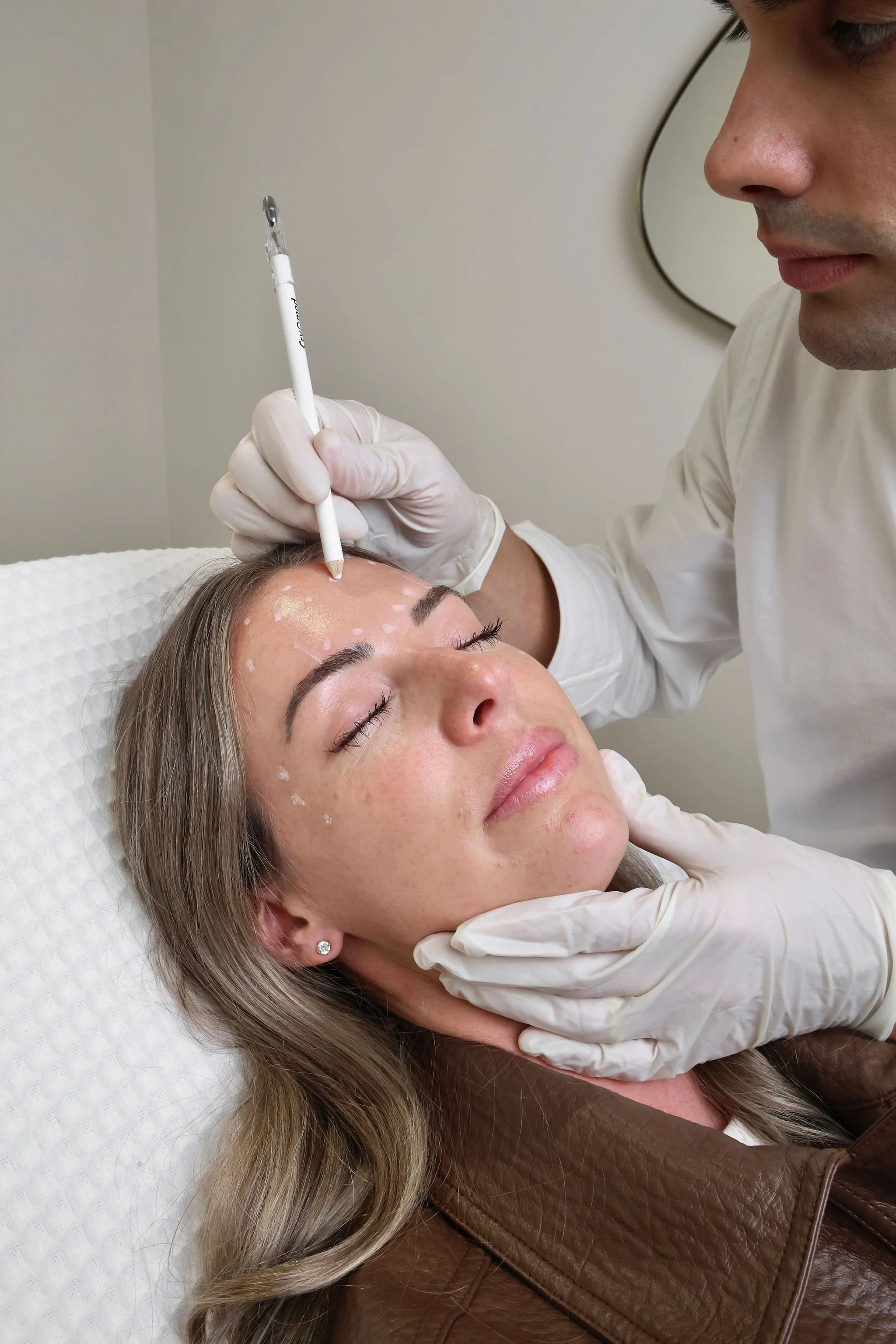 A woman lying on a medical bed with her eyes closed, receiving a cosmetic facial treatment from a healthcare professional. The professional is holding a pen-like tool and marking her face, wearing white gloves.