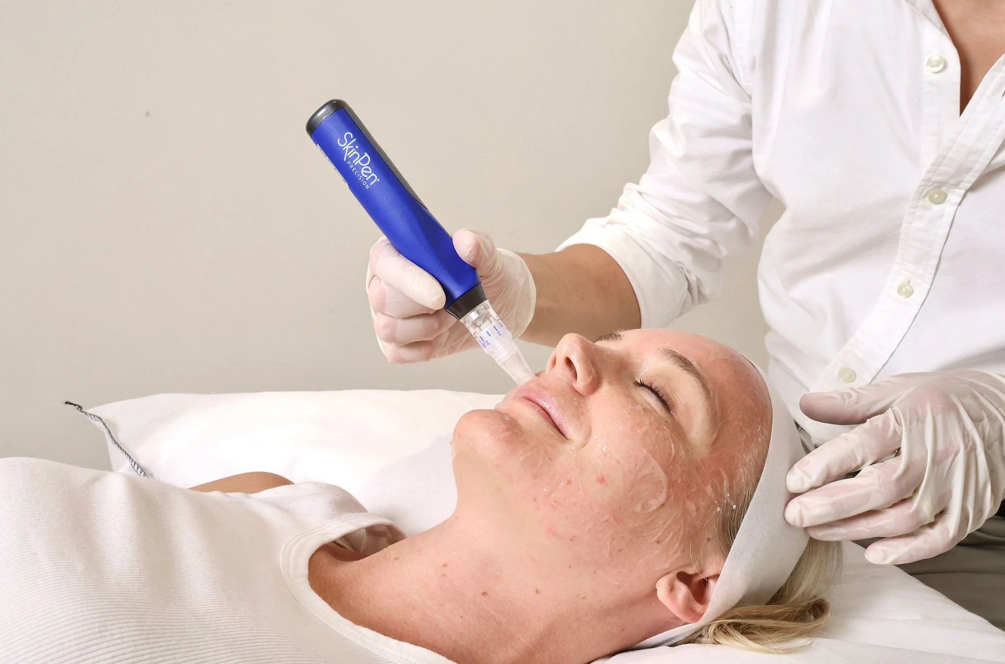 A woman is lying on her back with her eyes closed while a medical professional applies a facial treatment using a blue skincare device. The professional is wearing white gloves and a white button-up shirt.