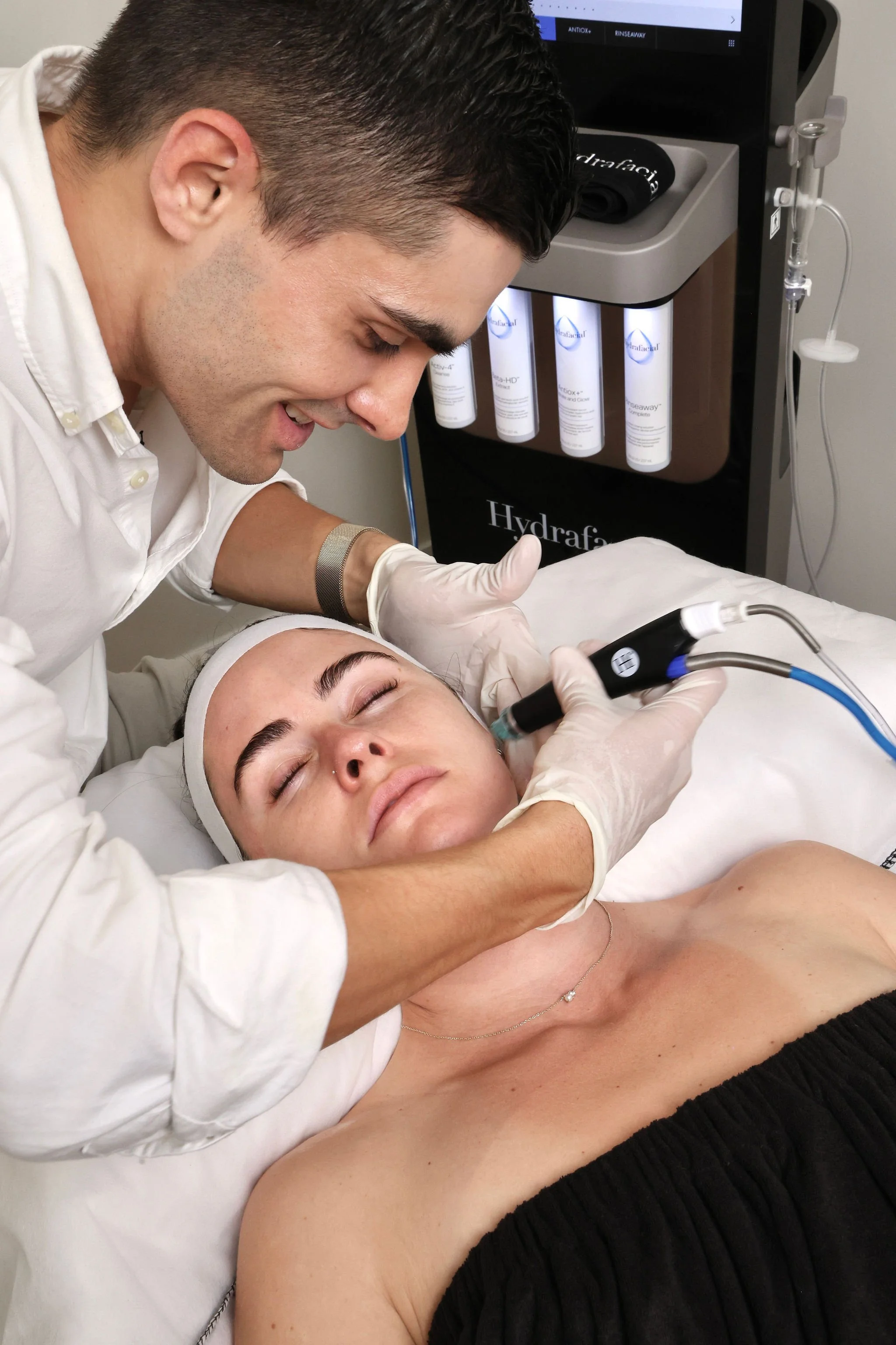 A healthcare professional performs a facial treatment with a small handheld device on a woman lying on a treatment bed, wearing a black strapless top and sleeveless, with her eyes closed. Medical equipment and skincare products are visible in the background.