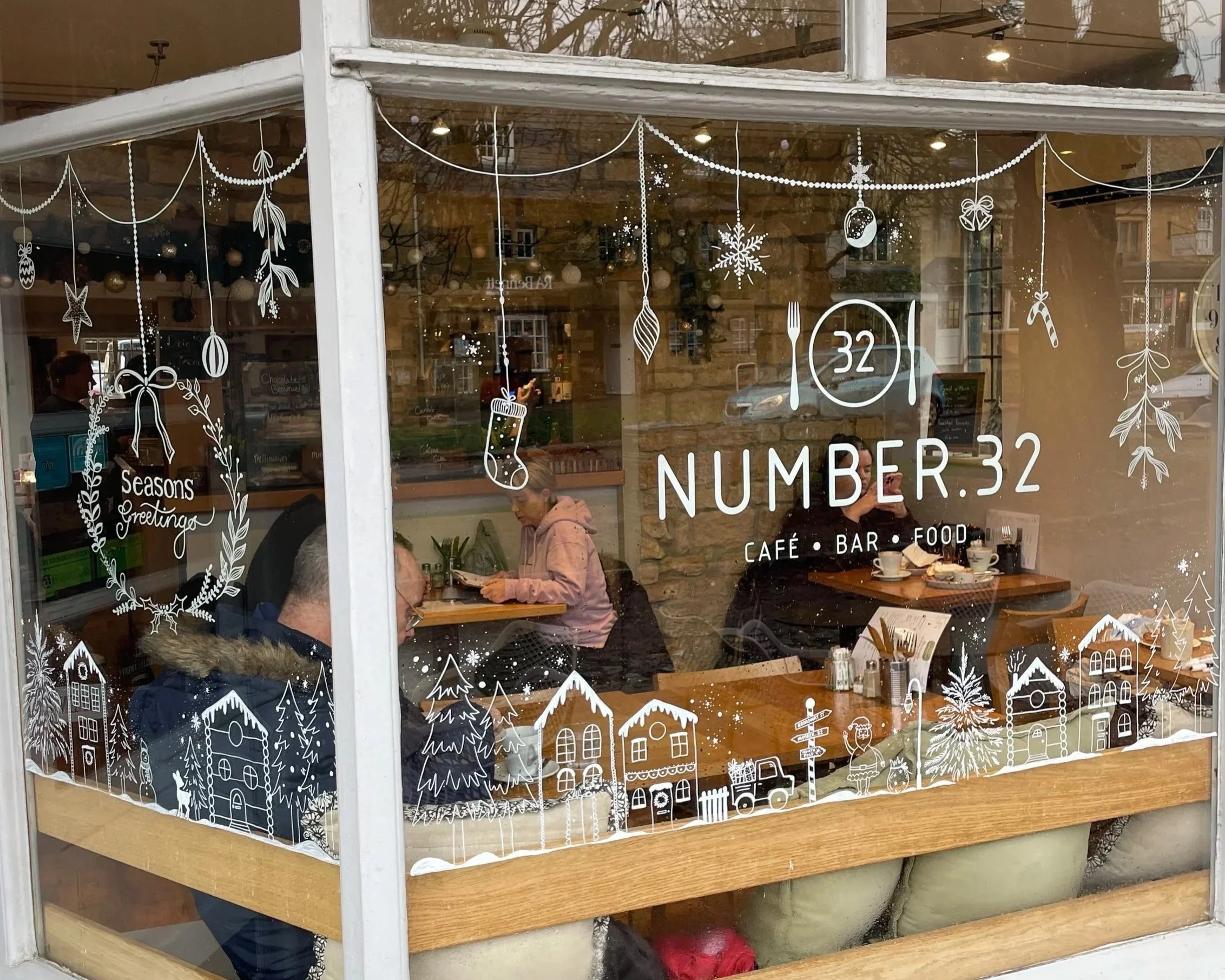 The window of a café called 'Number 32' decorated with white holiday-themed illustrations including snowflakes, trees, houses, and ornaments. Inside, patrons are seated at tables, and one person is looking at their phone.