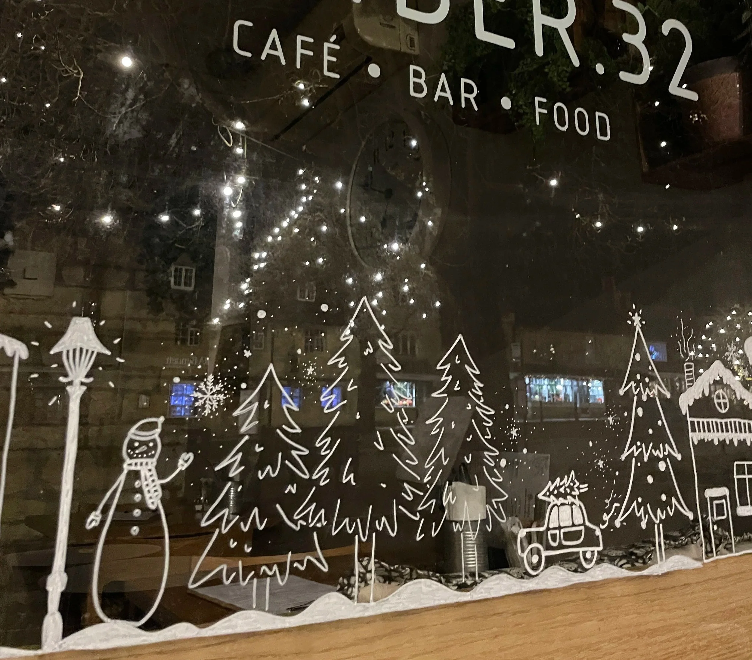 Decorative window with white line drawings of trees, snow, a snowman, and a car with a gift on top, with a night city reflection in the background. Sign reading 'CAFE BAR FOOD' at top.