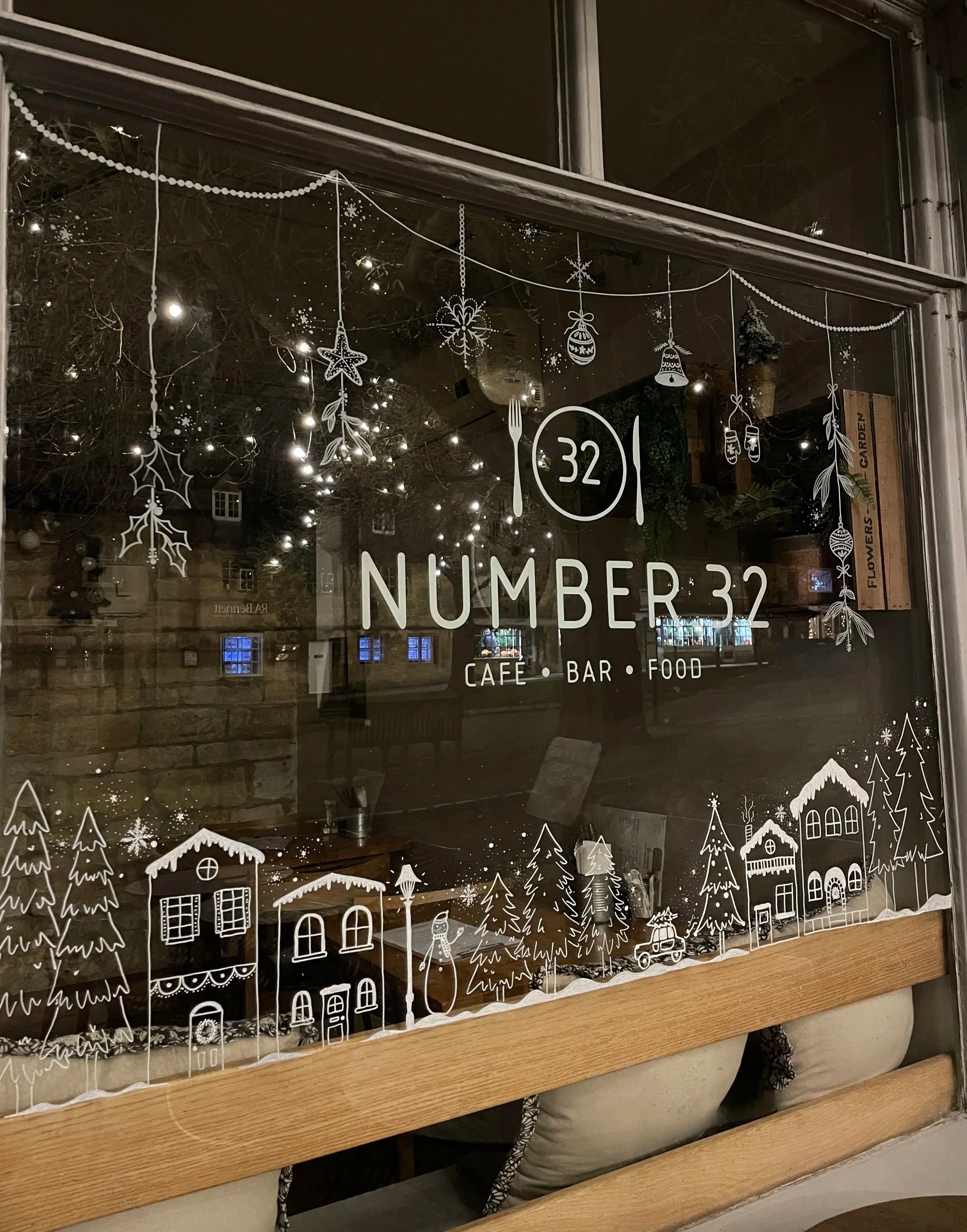 Decorative window display for Number 32 cafe, bar, and food, featuring white Christmas-themed illustrations of houses, trees, ornaments, snowflakes, and hanging decorations, with indoor string lights reflecting inside.