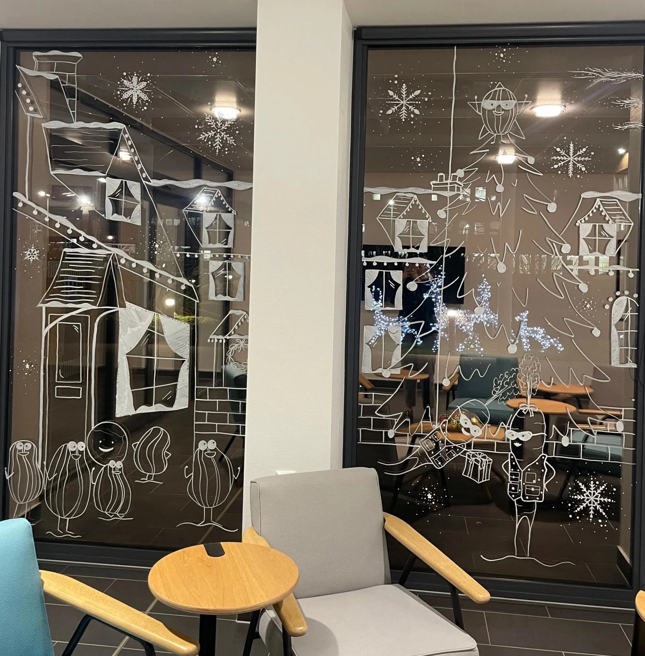 Decorative holiday window with white line drawings of a Christmas tree, a star, houses, a person with a Santa hat, and other festive elements, with snowflakes and stars.