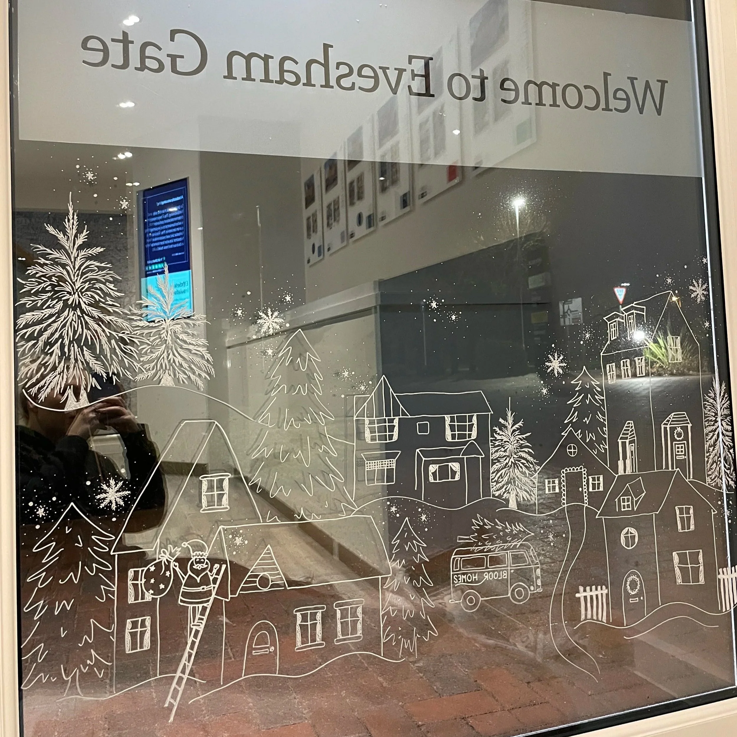 Decorative window with a white outline drawing of a winter village scene, including houses, trees, snowflakes, and a snowplow truck, with a reversed message at the top that reads "Welcome to Evesham Gate."