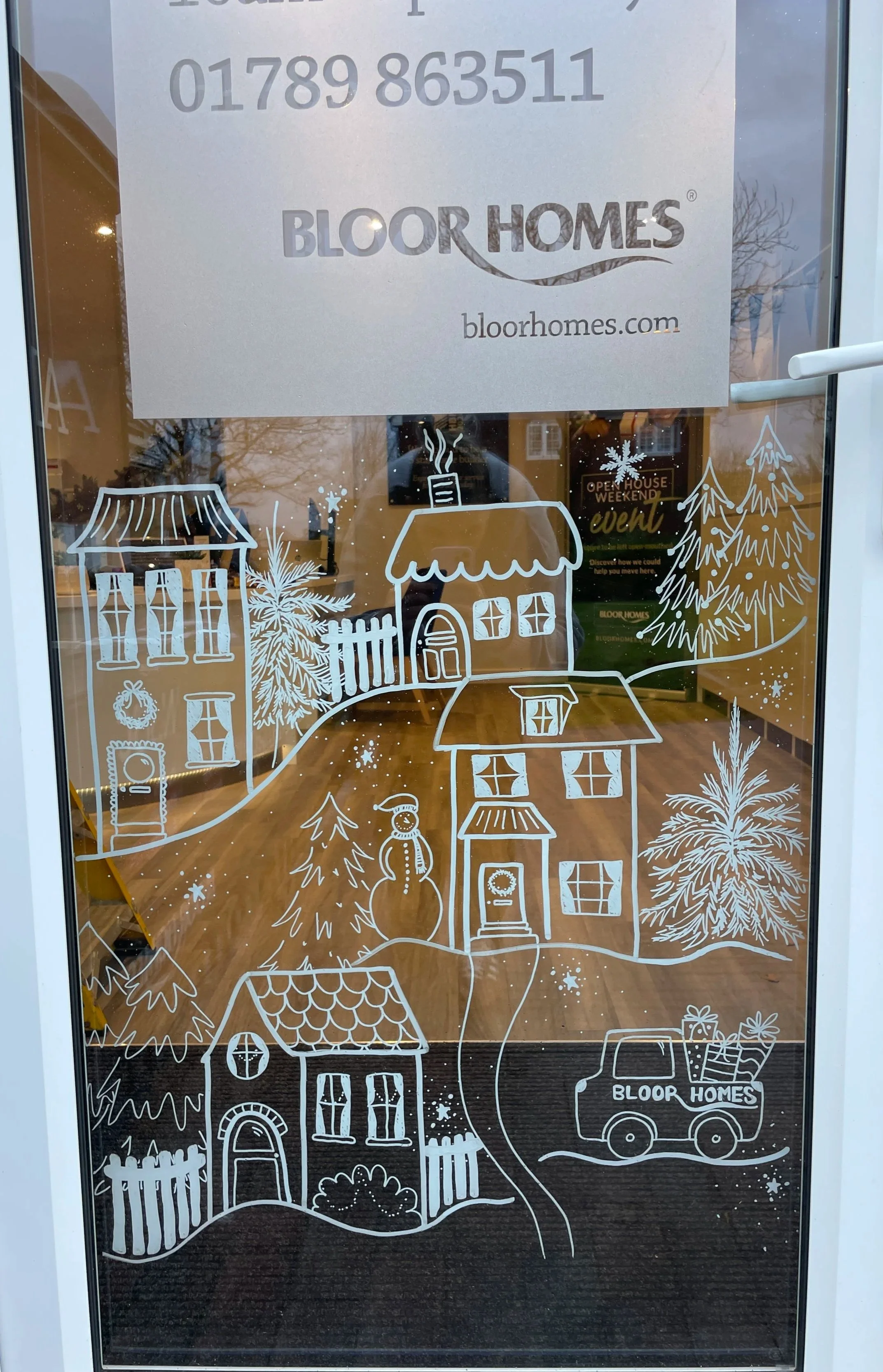 Decorative winter scene on a glass door with houses, trees, a snowman, and a truck, all in white outline, with a sign for Bloor Homes and a website address.