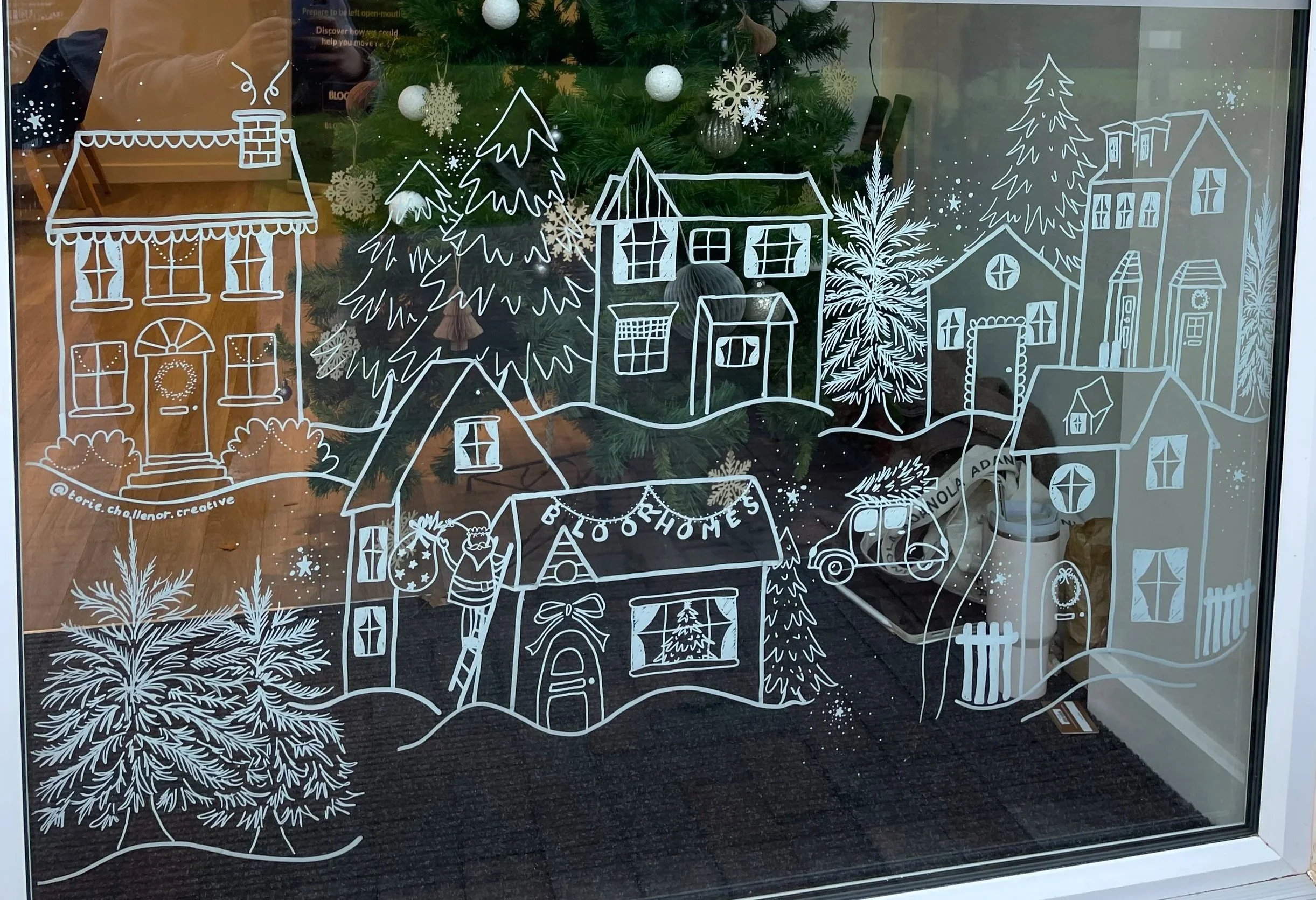 Decorative white drawing on glass window depicting a holiday scene with houses, trees, and a person hanging ornaments, with a Christmas tree visible inside behind the glass.