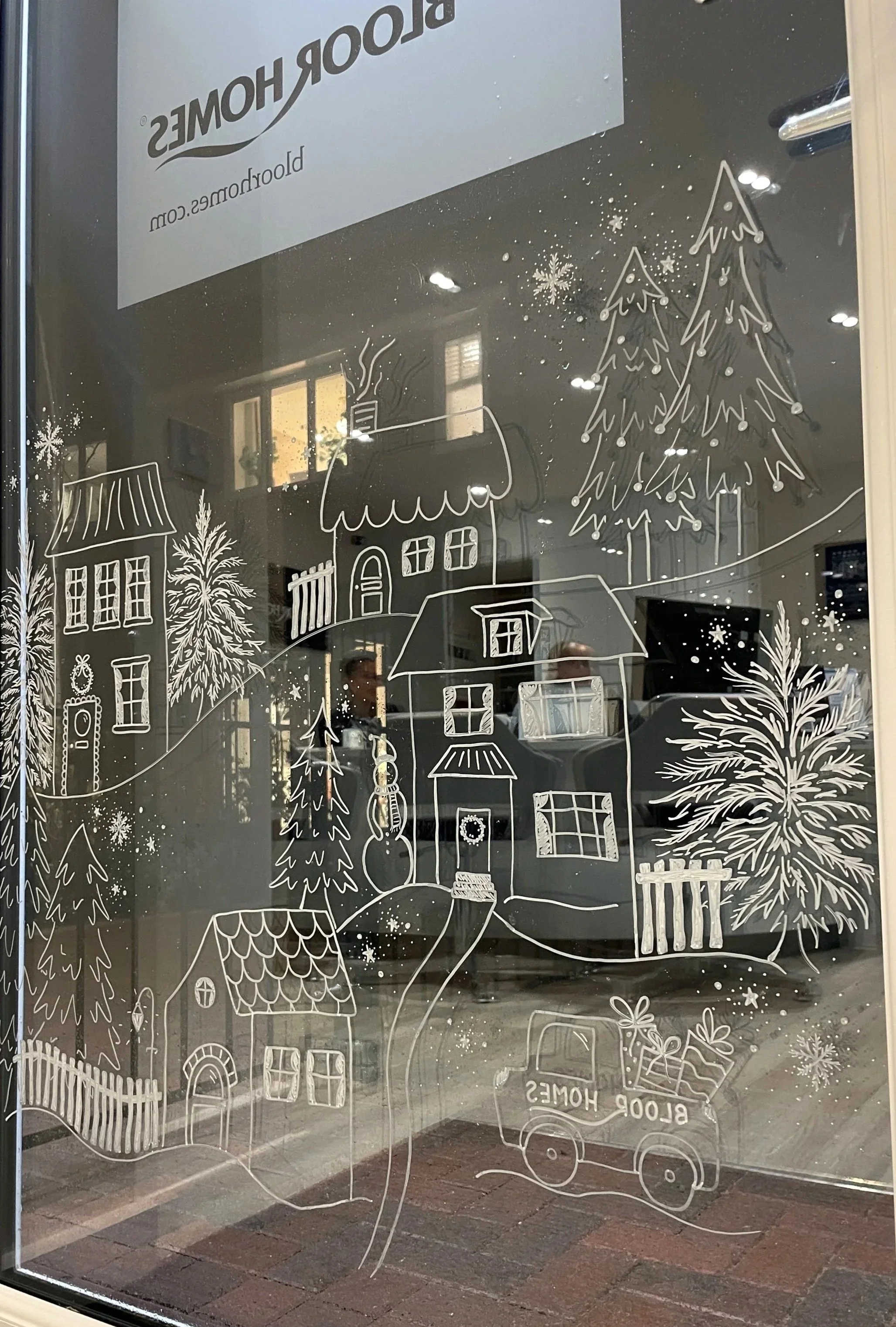 Decorative window illustration with a Christmas village scene featuring houses, trees, a snowman, and a truck filled with presents, all drawn in white lines on the glass.