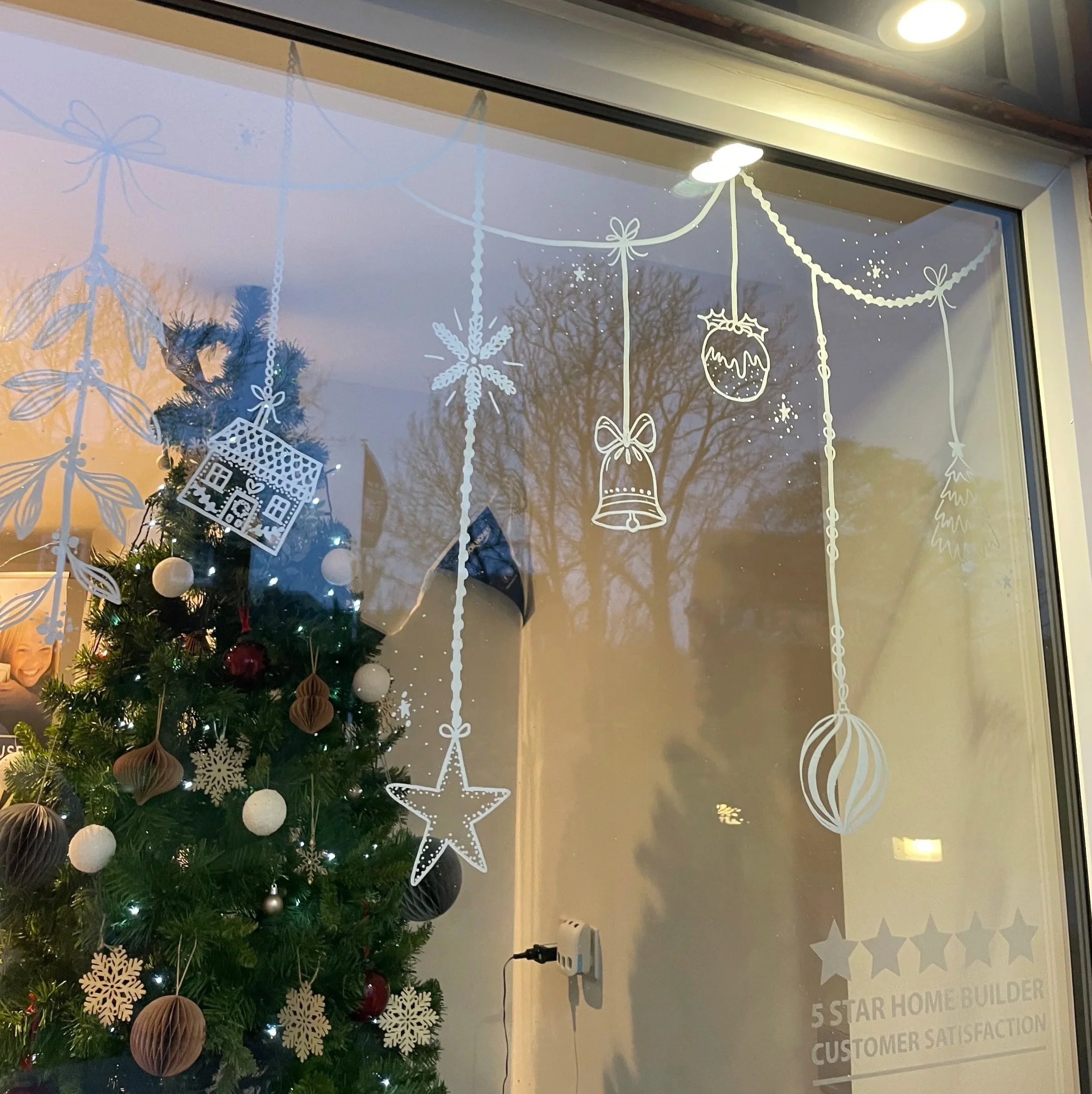 Christmas tree decorated with white and red ornaments inside a store windows with festive white holiday-themed decals including snowflakes, stars, bells, baubles, a house, and a candy, with a reflection of trees outside and a voucher for a five-star 