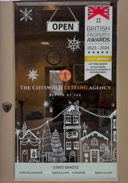Glass door with snowflake decals, an 'OPEN' sign, and a British Property Awards banner. The banner indicates the 2023-2024 gold winner for letting agent in southern Gloucestershire and Oxfordshire. The door has a handle on the left side, and the inte