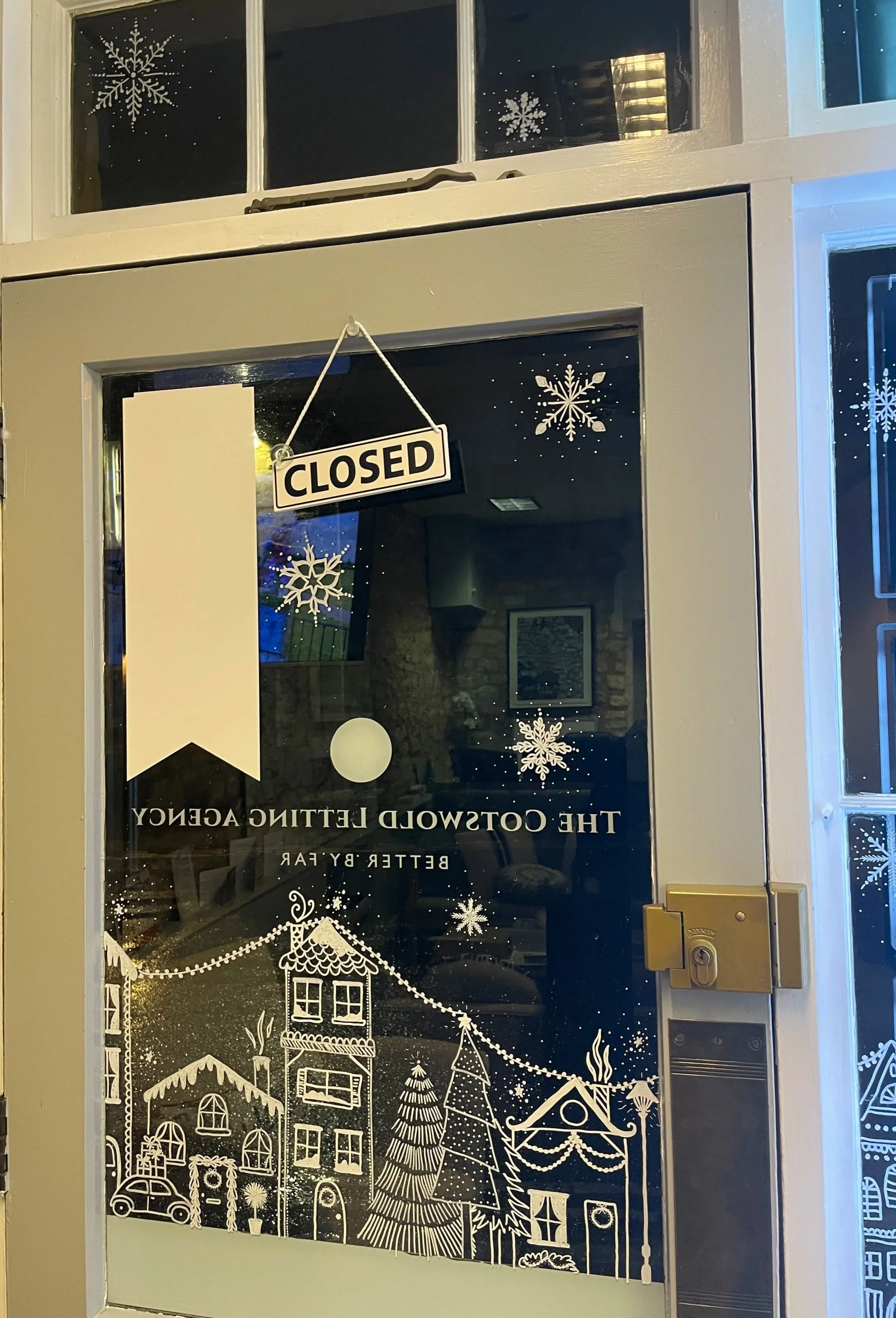Decorated glass door with white winter-themed illustrations of houses, trees, snowflakes, and string lights, with a "CLOSED" sign hanging on the door.