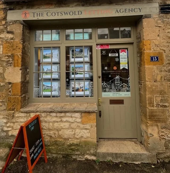 The front entrance of The Cotswold Lettering Agency, housed in an old stone building with a glass window and door. The window displays property listings, and there is a sign on the door indicating that the office is open. A small blackboard sign outs