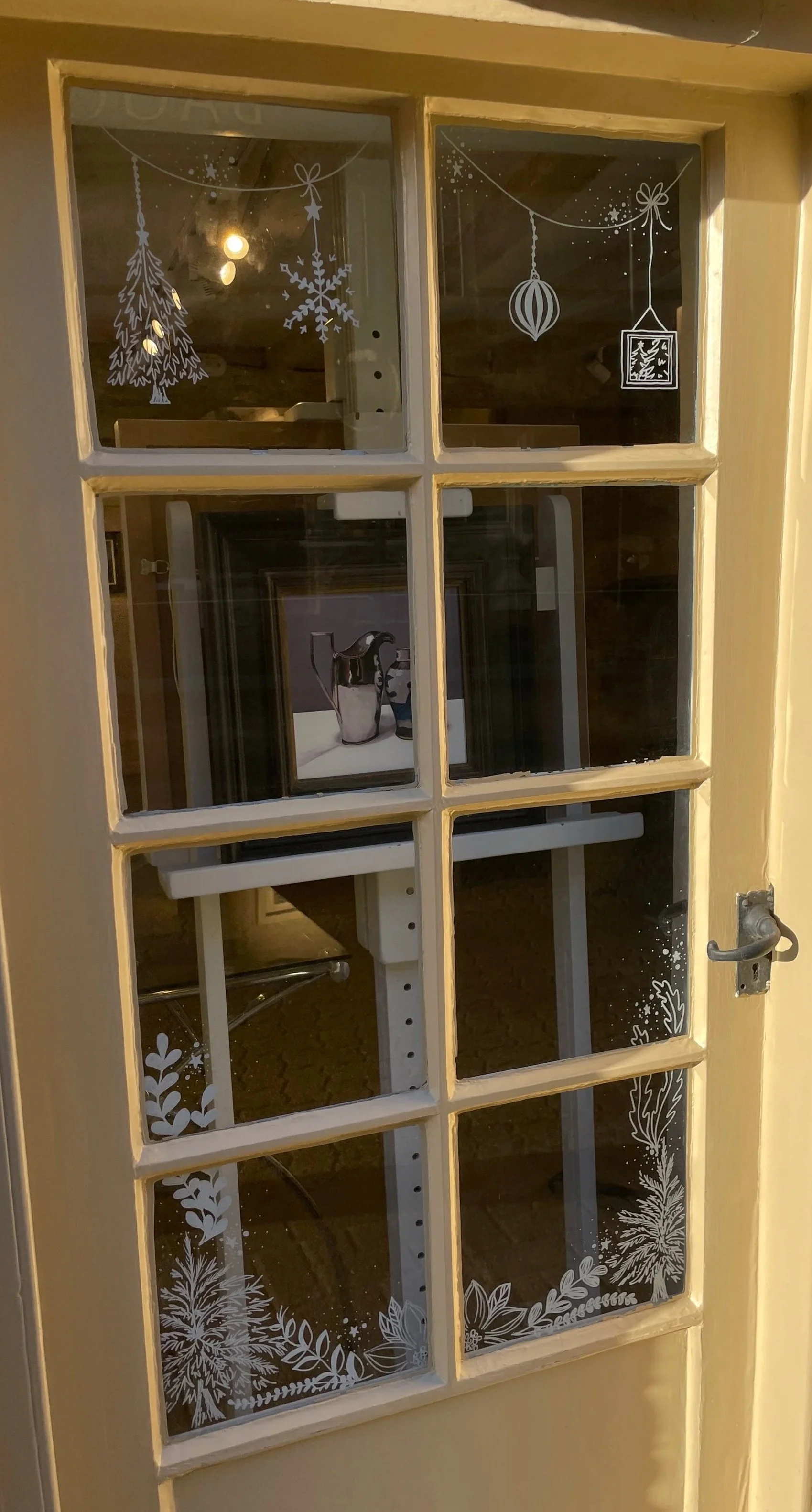 Decorative etched winter holiday designs on a glass door window, featuring a Christmas tree, snowflake, hanging ornament, gift box, and various leaves and branches.