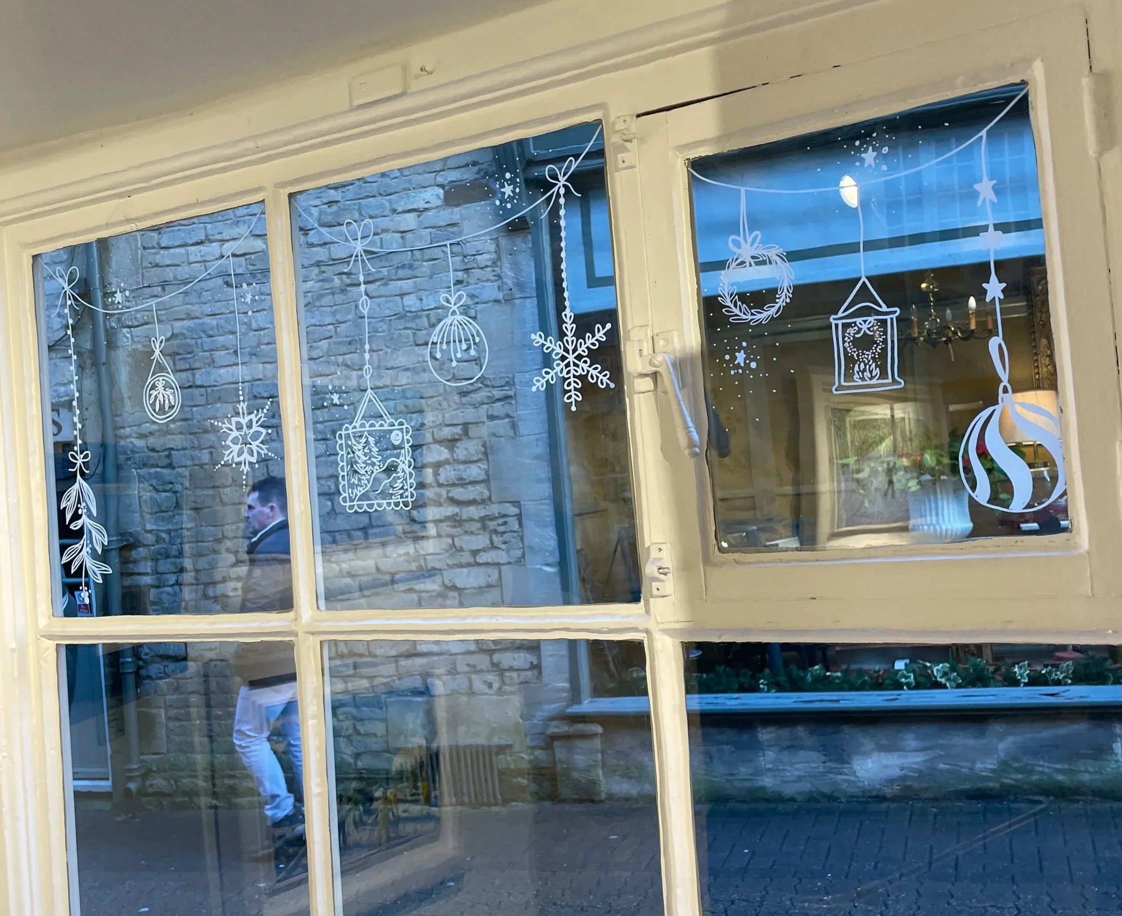 Decorative Christmas window clings featuring snowflakes, ornaments, a wreath, a lantern, and other holiday designs on a window pane, with a brick building and a person reflected in the glass.