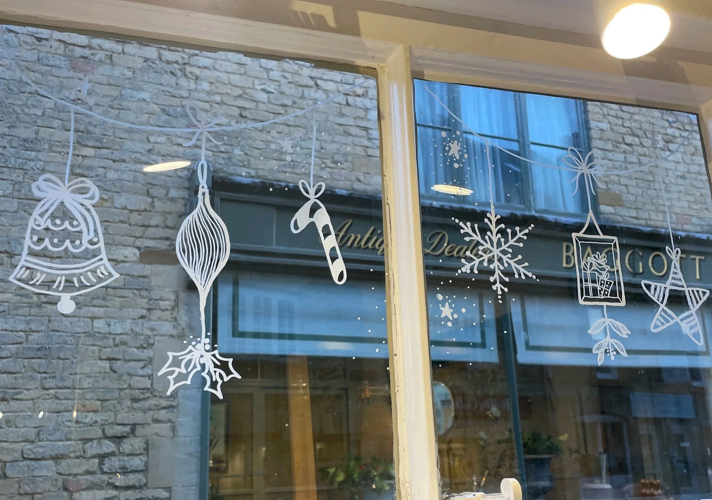 Decorative white Christmas ornaments, including a bell, a teardrop-shaped ornament, a candy cane, a sprig of holly, a snowflake, a gift box, and a star, hanging on a window.