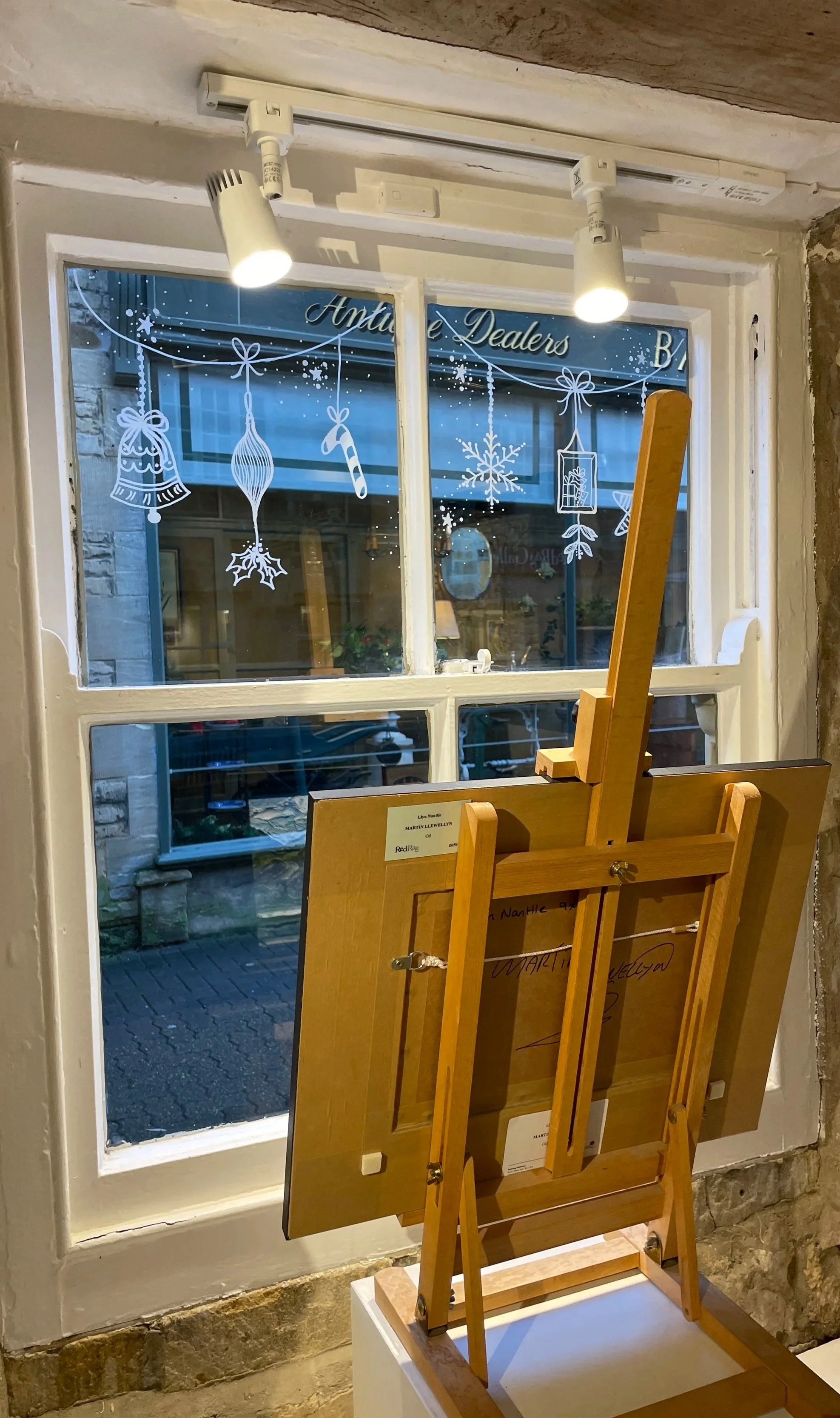 View of a painting easel placed in front of a window. The window has holiday-themed white stickers including bells, ornaments, a candy cane, a snowflake, and a gift box. The scene outside the window shows a street with buildings and parked cars.