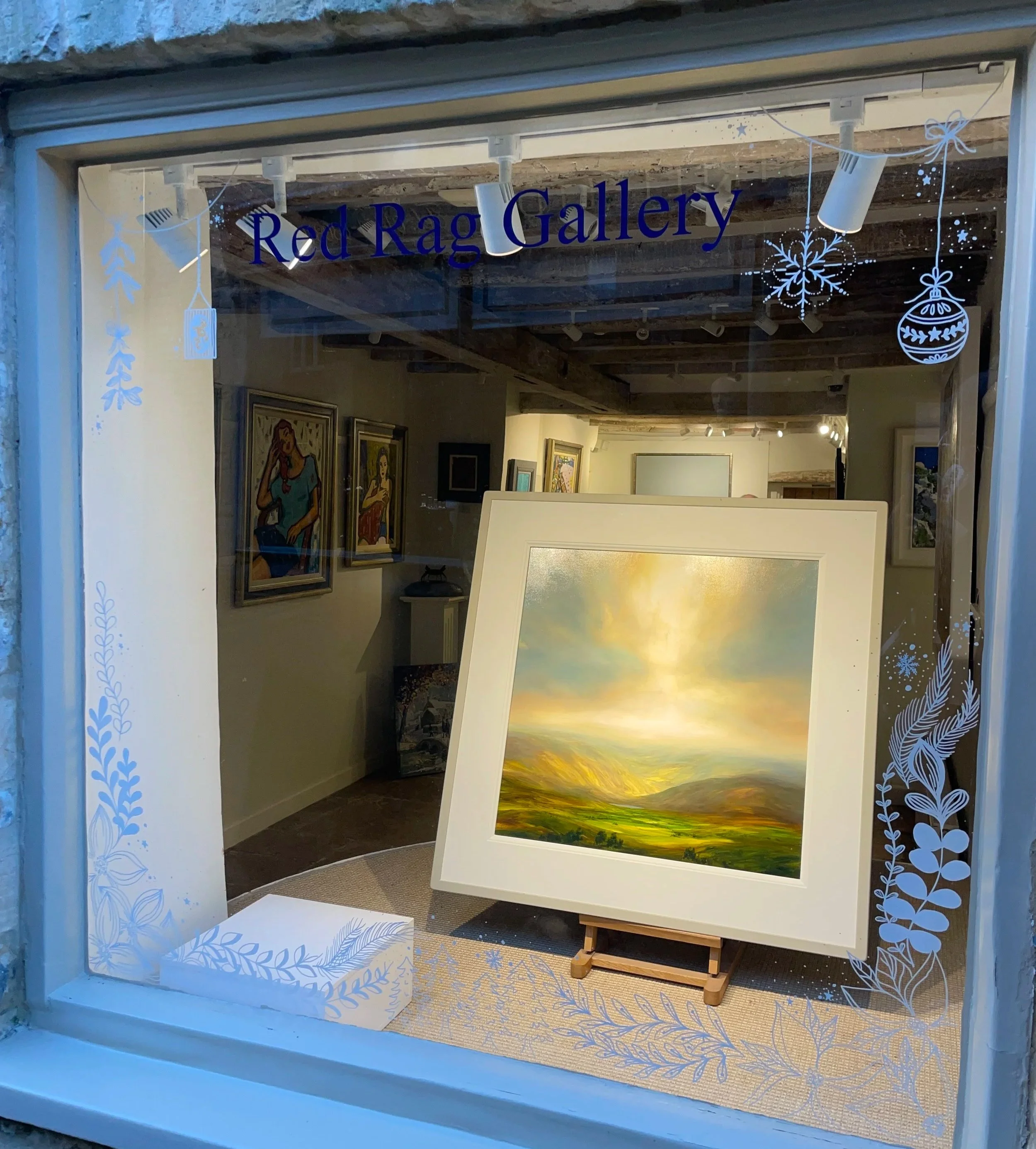 Art gallery window display with landscape painting on an easel, featuring mountains, green fields, and a glowing sky. Inside, framed artworks are visible on the walls, with the gallery name 'Red Rag Gallery' written above in black letters.