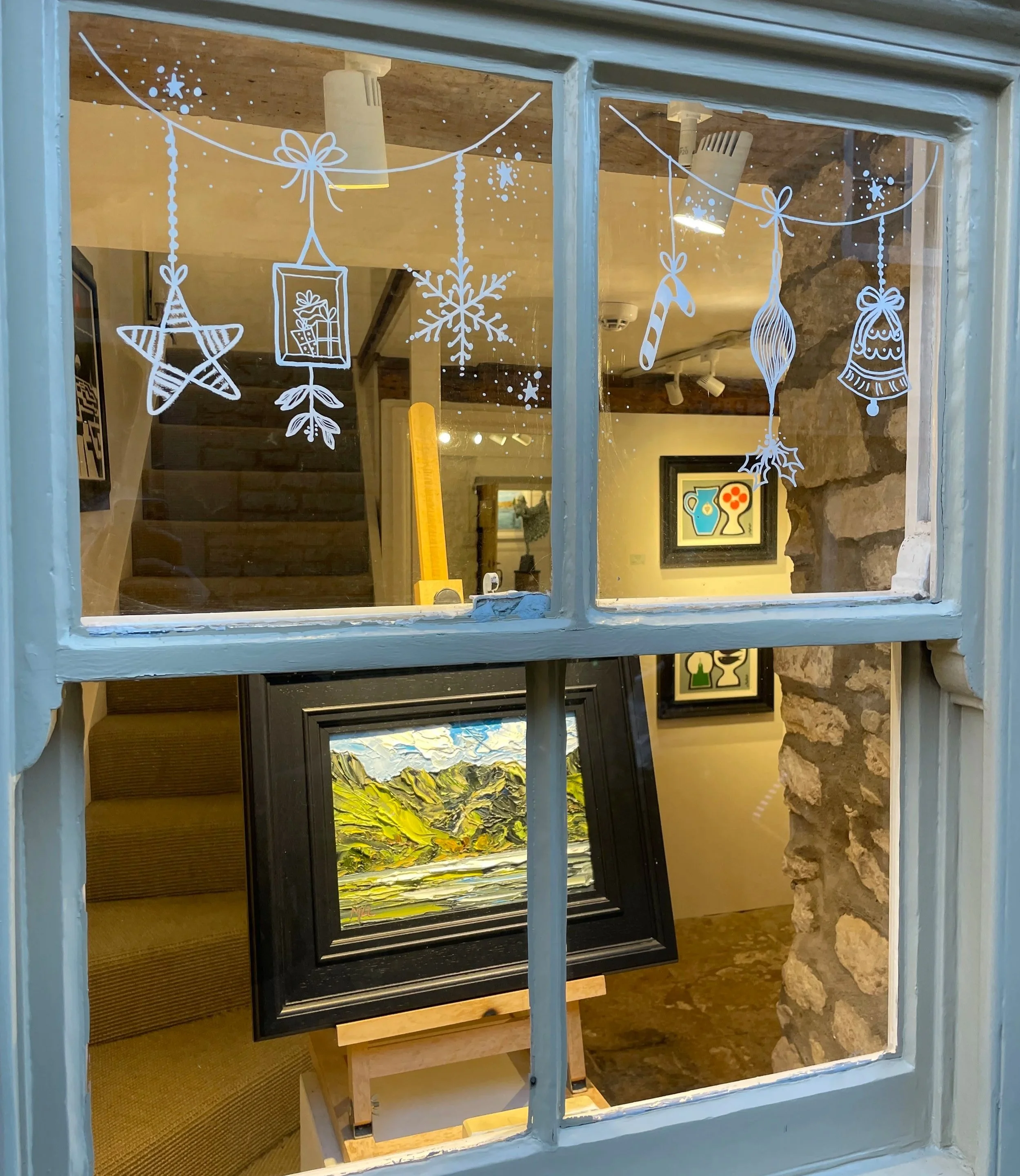 View through a window showing an art gallery interior with holiday decorations, including white string art of stars, snowflakes, a gift, a candy cane, and ornaments. Inside, a framed landscape painting is on an easel, and another framed artwork is on