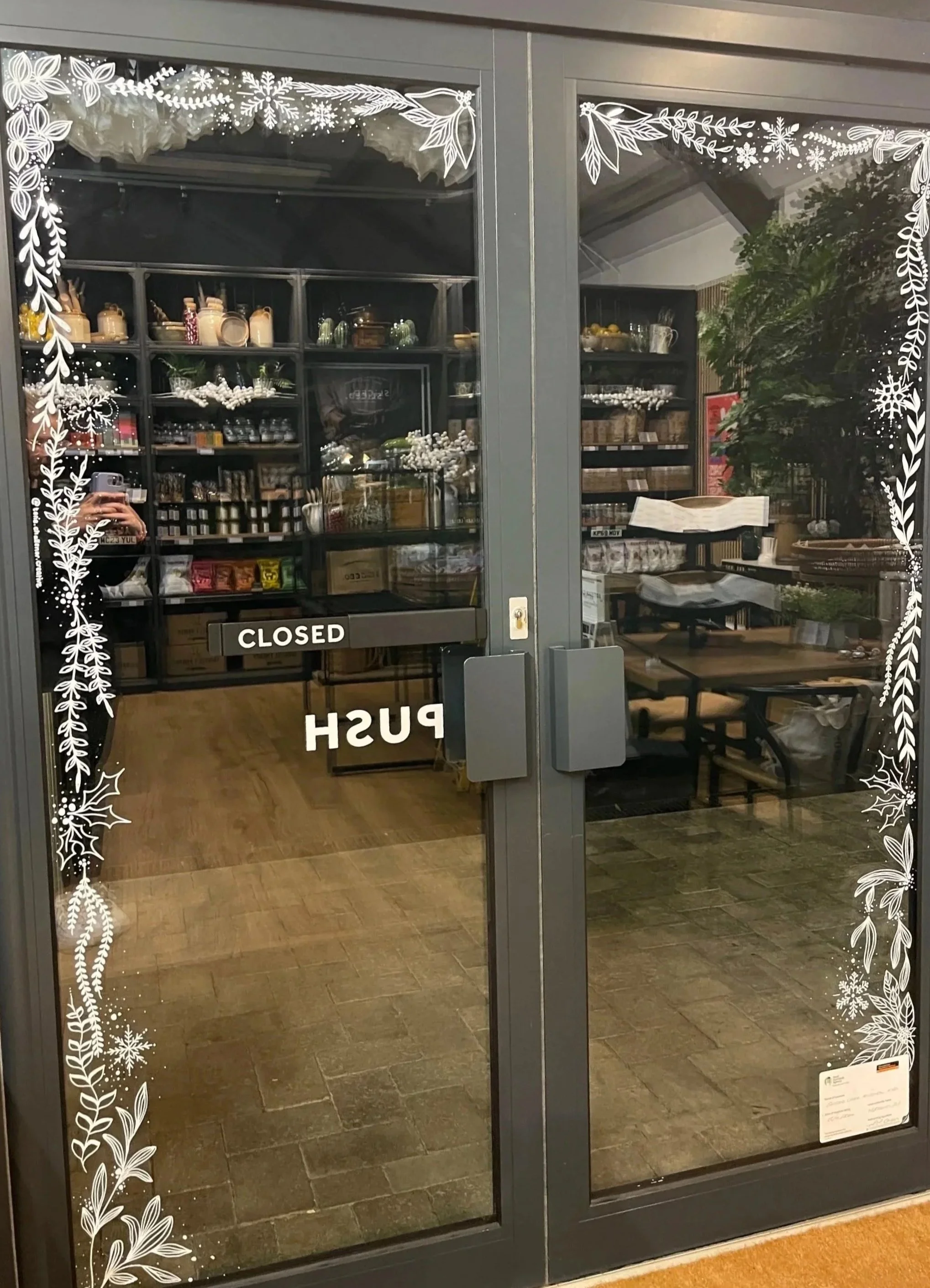 Glass door with holiday snowflake and foliage window art, showing a closed shop interior with shelves of goods and a table and chairs inside.