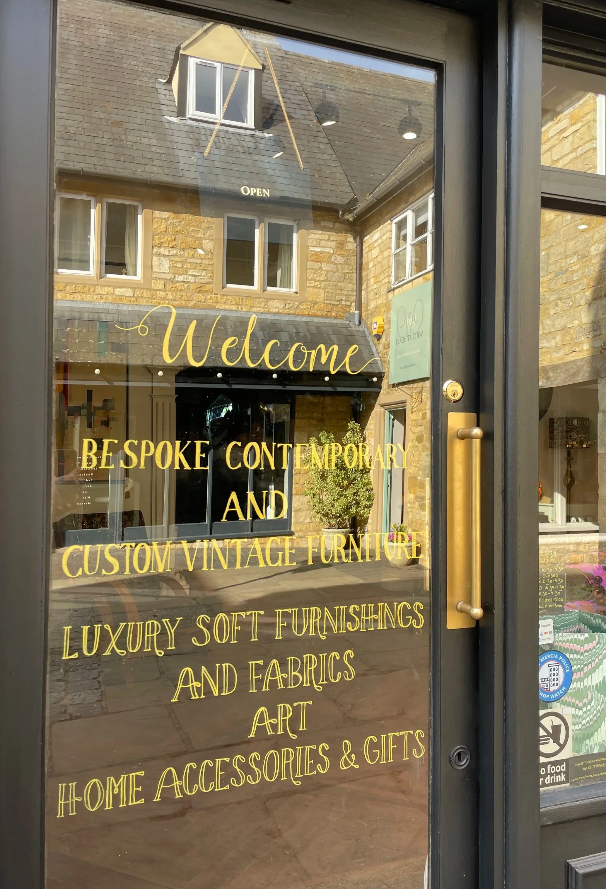 Glass door with yellow text welcoming visitors to a store that sells bespoke contemporary and vintage furniture, soft furnishings, fabrics, art, home accessories, and gifts. The store is in a brick building with windows, and there is a small awning a