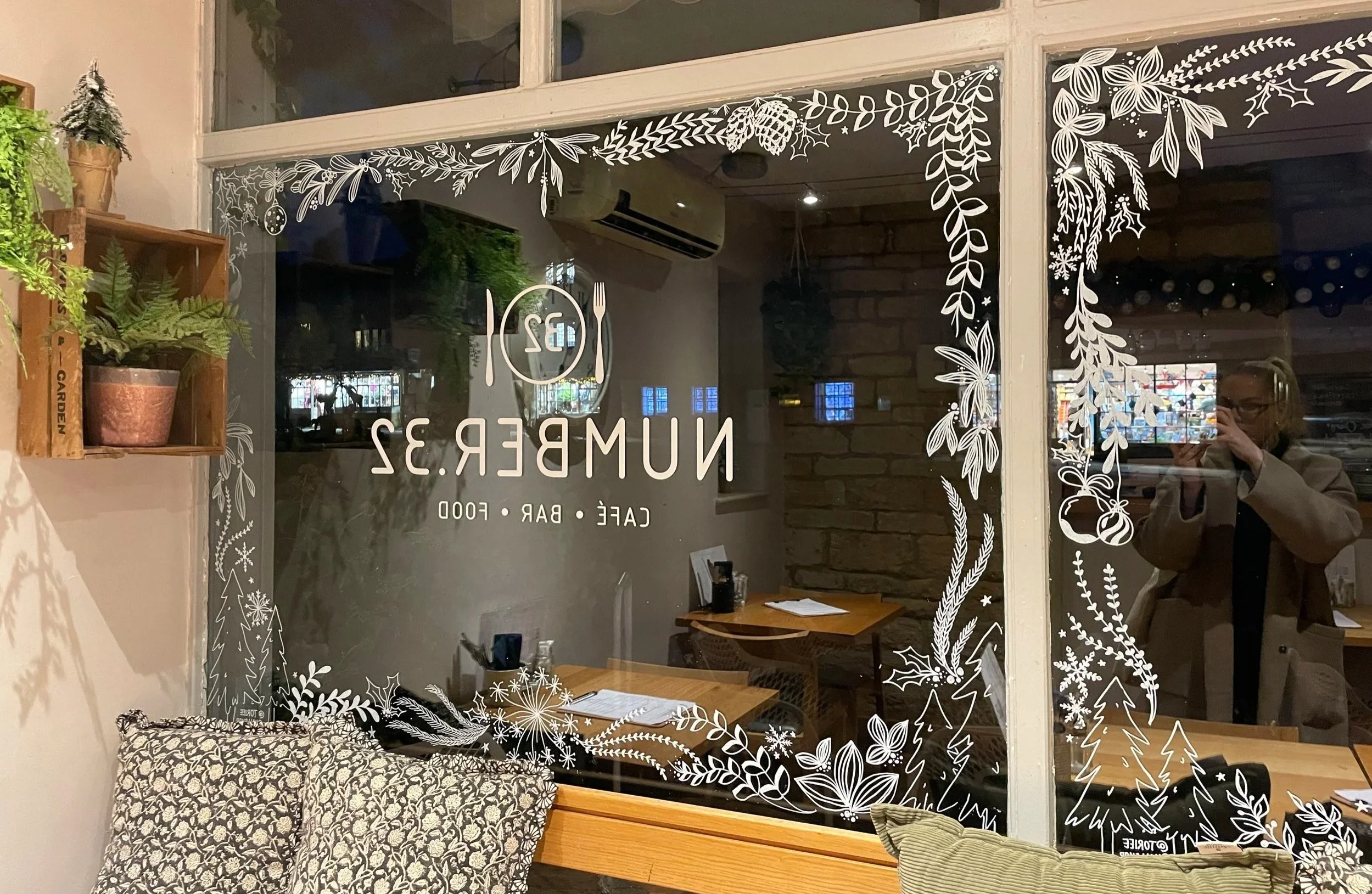 Interior of a cafe with a large glass window decorated with white holiday-themed illustrations including trees, leaves, and ornaments. Inside, there are wooden tables with menus, a brick wall, and hanging plants. A person is seen taking a photo in th