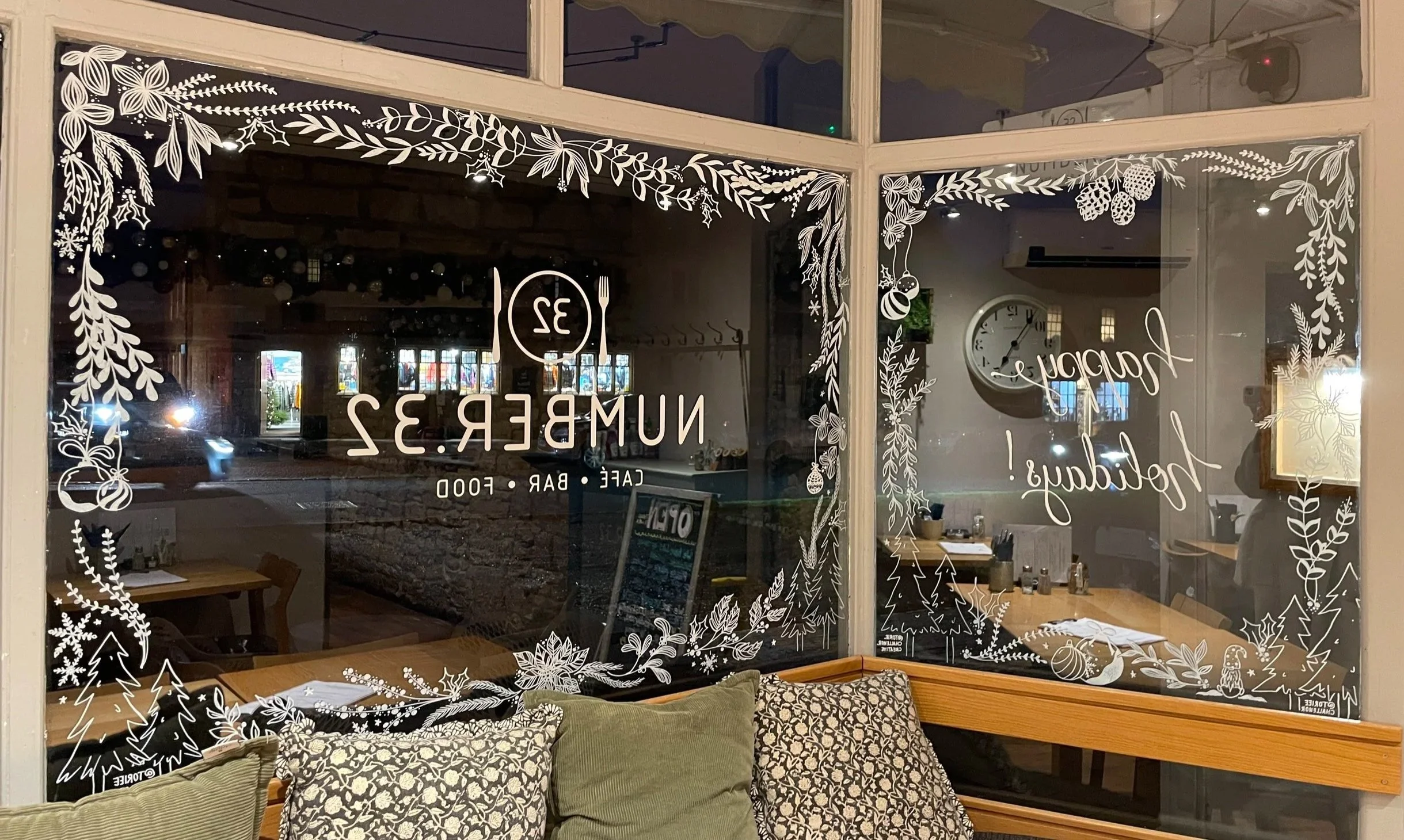 Decorative window with white holiday-themed floral and pinecone designs, with text indicating "Numb3r 32 Cafe Bar Food" and a slogan "Happiest Holiday!" in a restaurant setting at night.