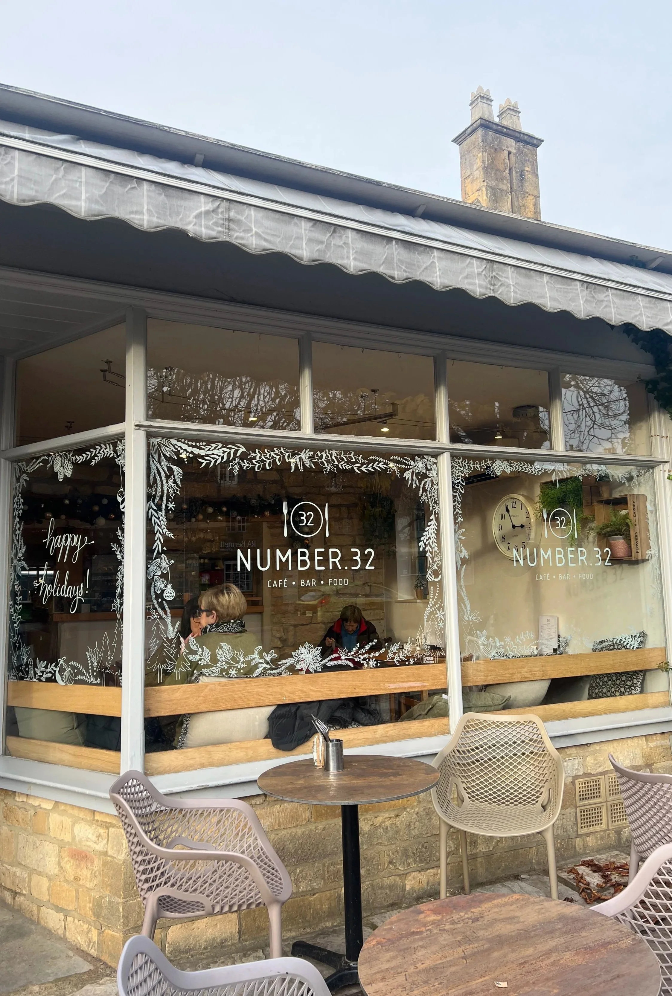 Exterior view of a cafe with large glass windows decorated with white holiday-themed designs. The cafe is named 'Number 32' and offers café, bar, and food services. There are outdoor tables and chairs in front of the cafe, and inside, a few people ar