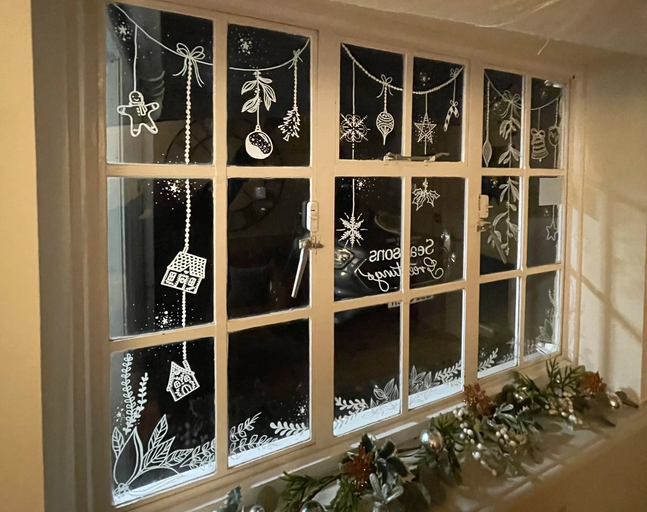 Decorative white paper snowflakes and holiday motifs hanging on a window, with Christmas garland and ornaments on the windowsill.