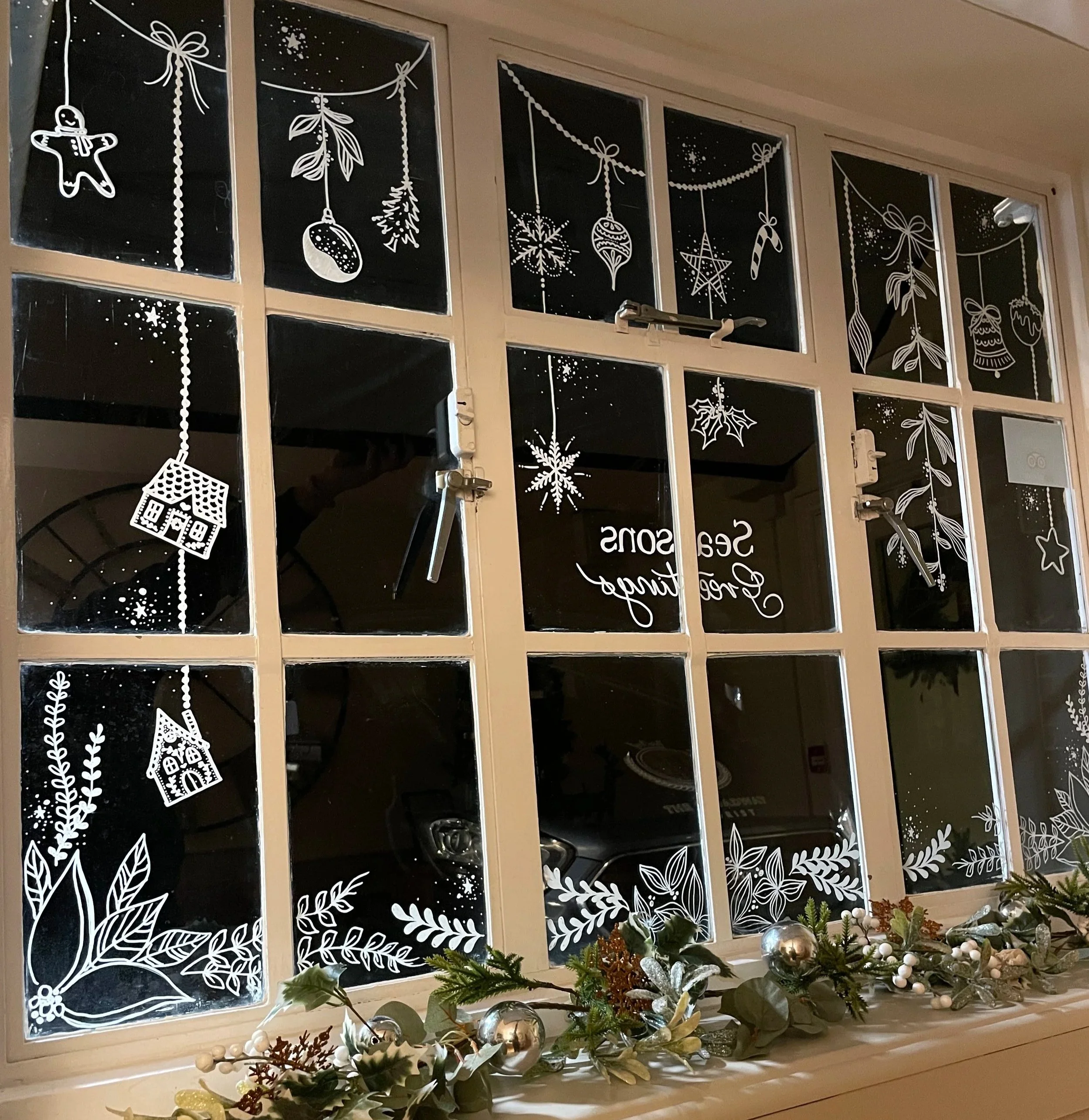 Decorated Christmas window with white holiday-themed drawings including gingerbread men, ornaments, snowflakes, holly, stars, and holly berries, with a festive garland of greenery and silver baubles along the window sill.