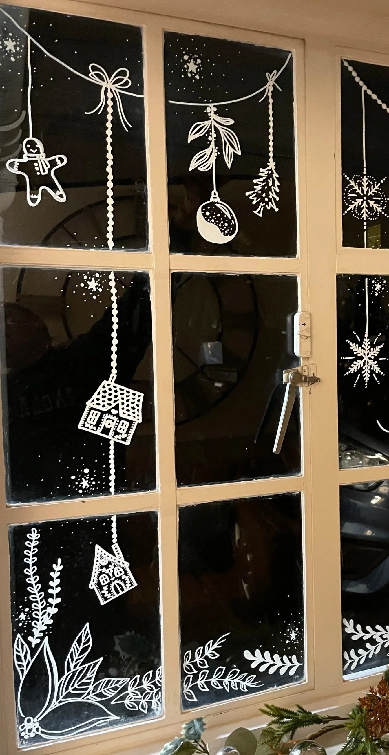 White Christmas-themed window decorations with snowflakes, houses, gingerbread man, pine branch, and hanging ornaments on a glass window.