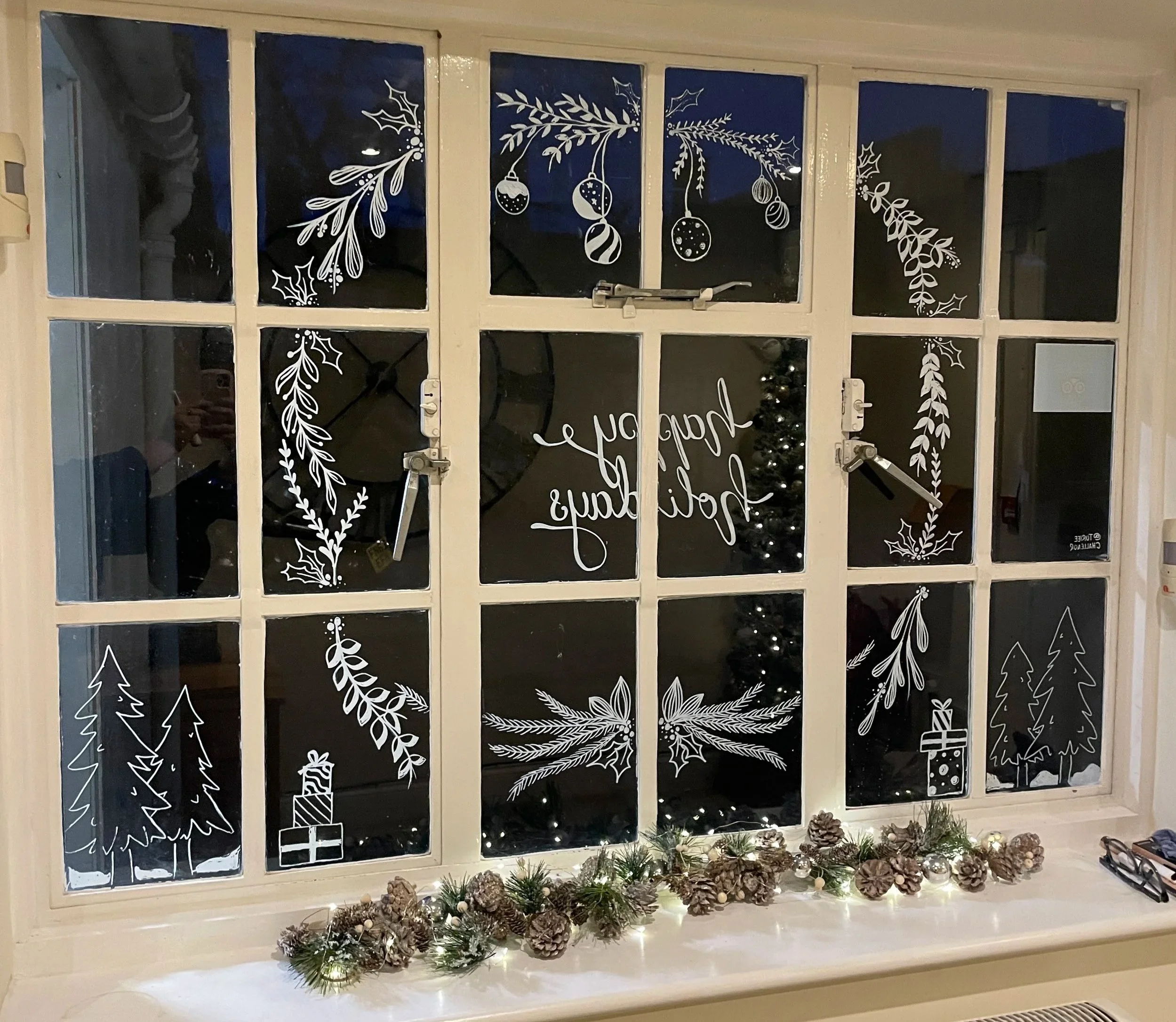 Decorated window with white winter-themed illustrations including pine trees, holly, ornaments, and a gift box. There is a Christmas tree with lights inside and a pine garland with pinecones and lights on the windowsill. The window has a message that