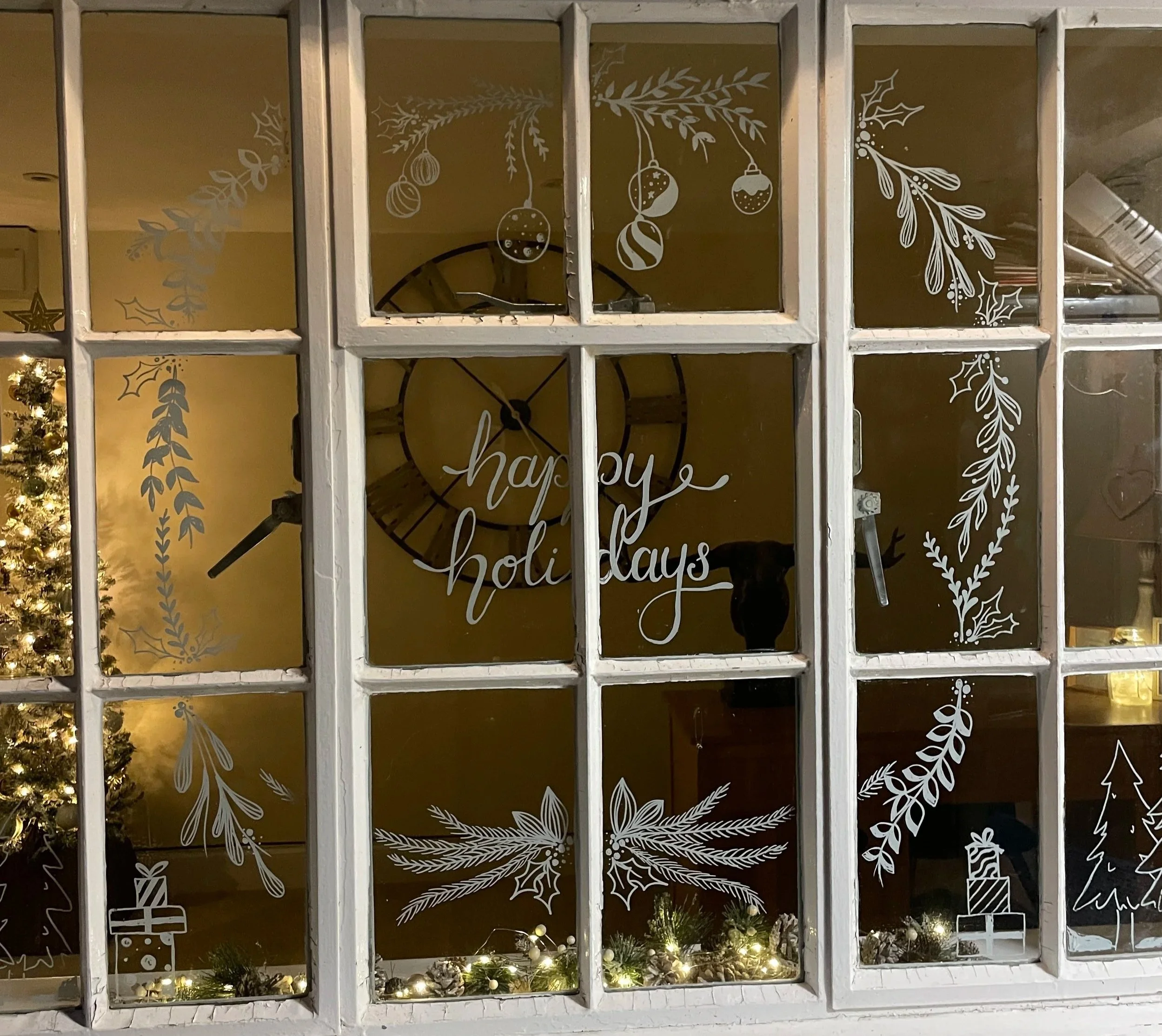 Decorated window with holiday and Christmas-themed white decals, including a Christmas tree, presents, candles, holly, and pine branches, with a "happy holidays" sign and a large clock in the background, along with twinkling string lights at the bott