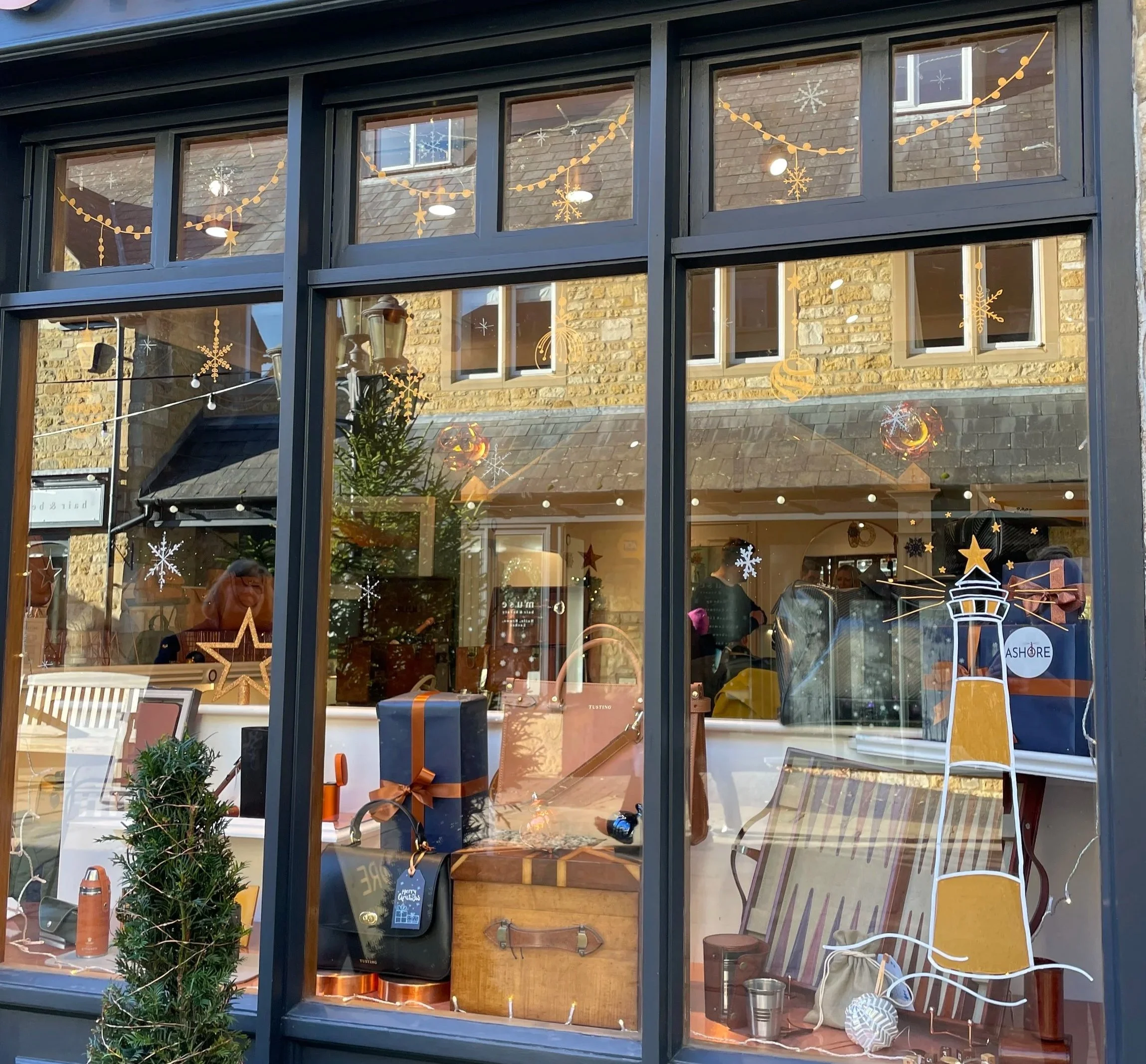 Store window decorated with Christmas-themed decals, including snowflakes, stars, and a lighthouse graphic. Inside, holiday gift items, a small Christmas tree, and shoppers are visible.