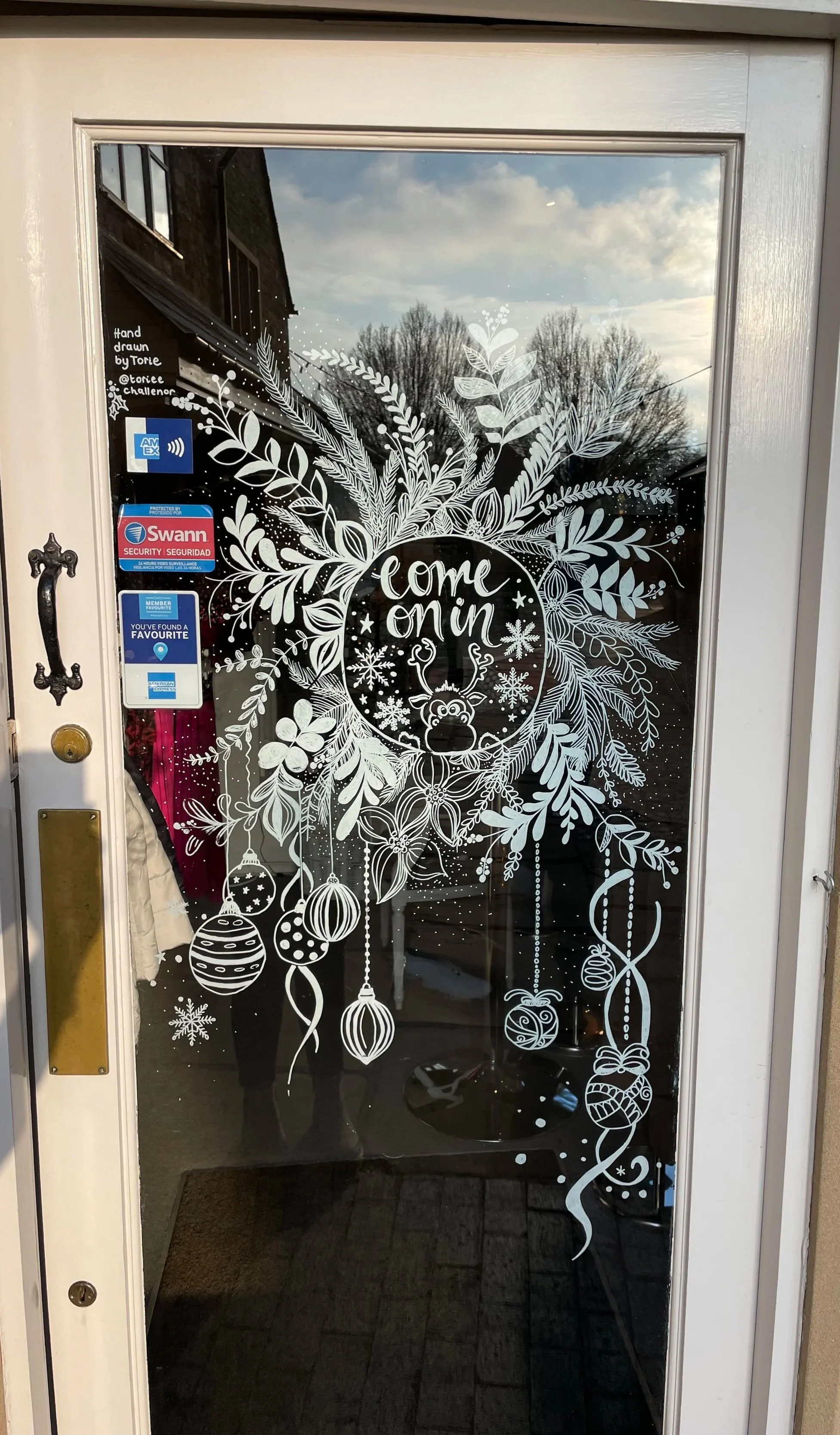 Decorated glass door with Christmas-themed white decals, including pine branches, ornaments, snowflakes, and a reindeer, with the words "come on in" in the center.