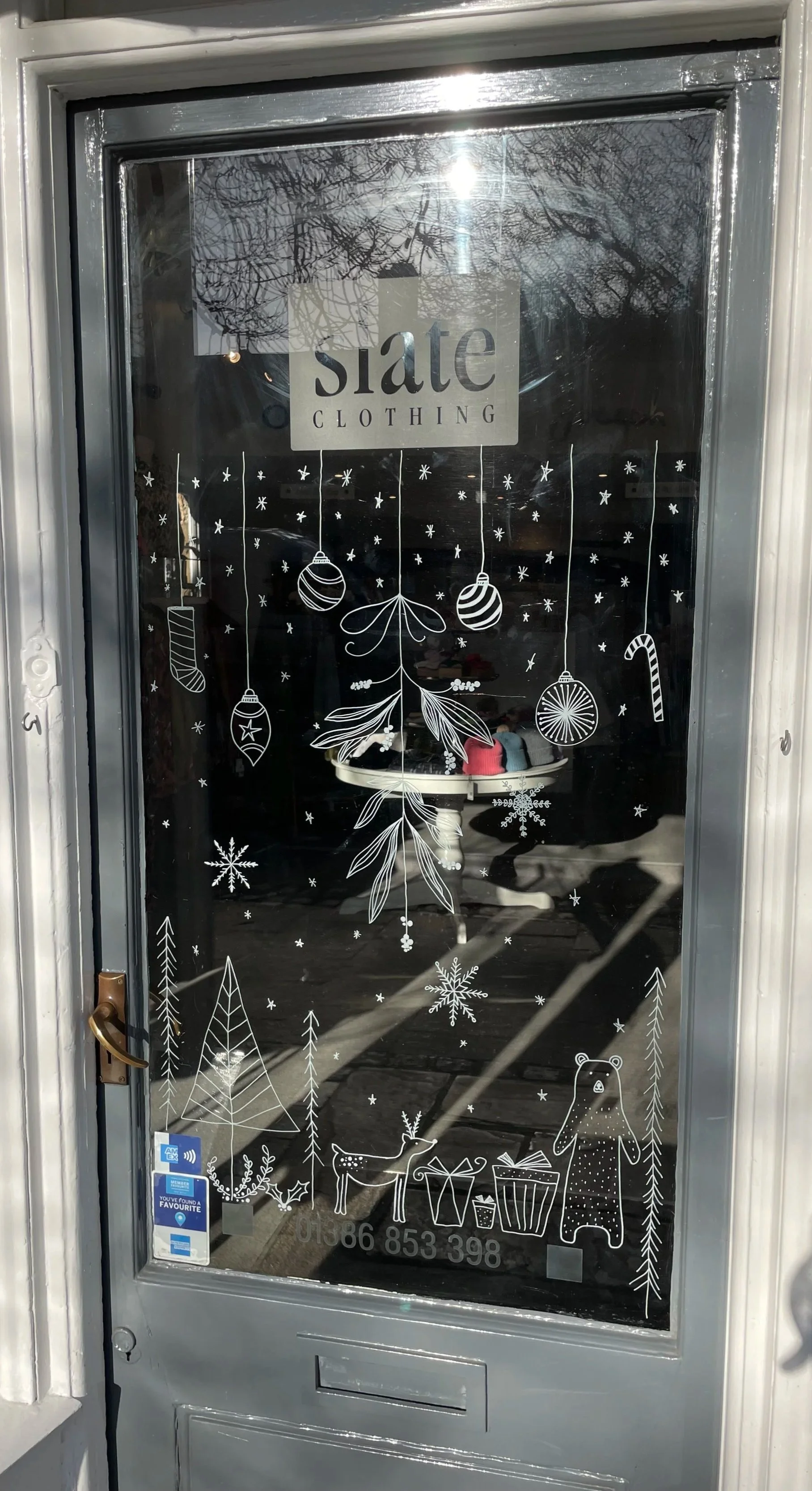 Decorative holiday illustrations on a glass door, including a reindeer, a bear, presents, snowflakes, Christmas ornaments, trees, holly, leaves, stockings, and candy canes, with the store signage for State Clothing at the top.