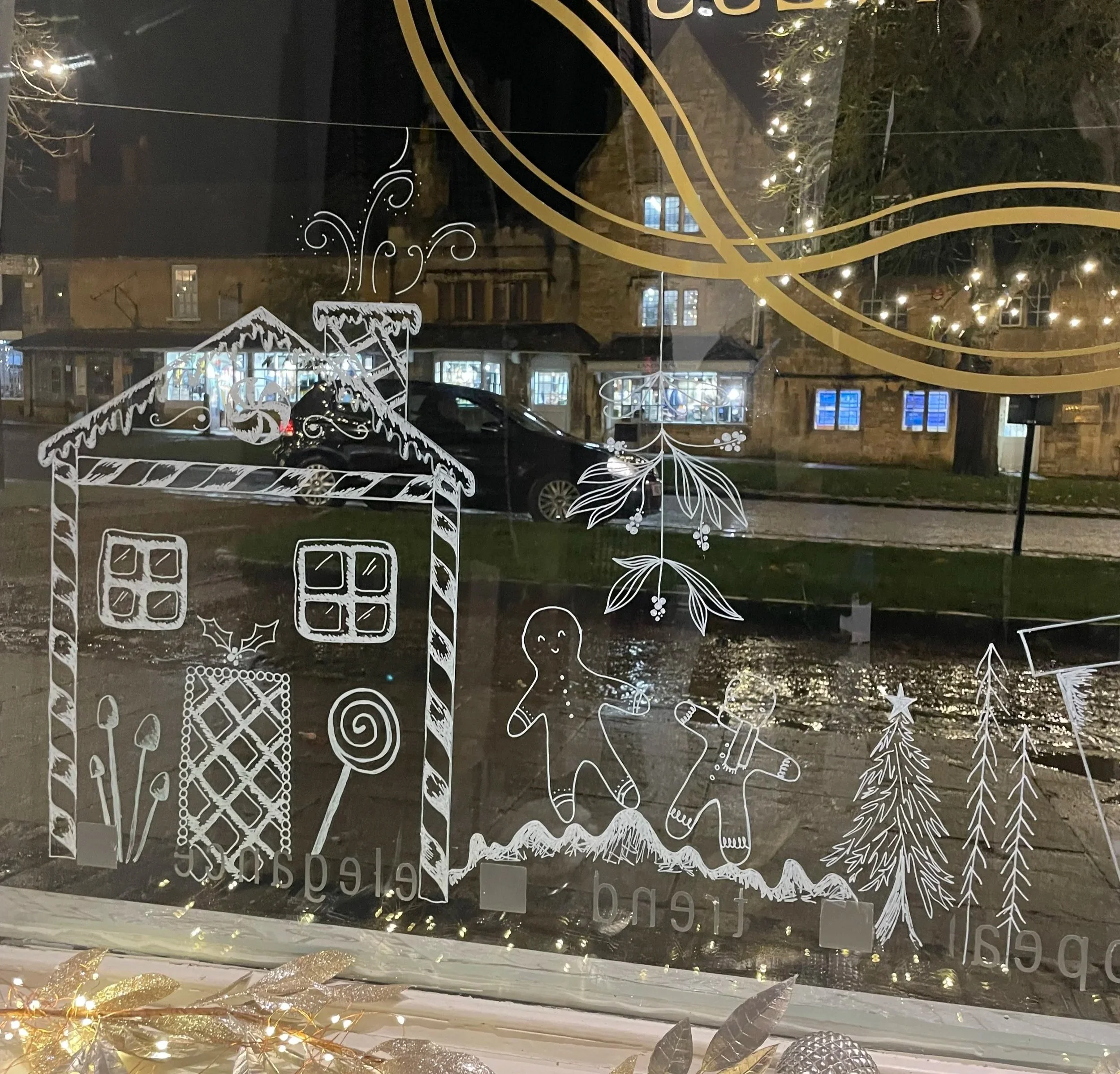 Decorative Christmas window with illustrations of a gingerbread house, gingerbread figures, Christmas trees, holly leaves, and candy canes, with a reflection of a street scene outside showing houses, parked cars, and illuminated windows at night.
