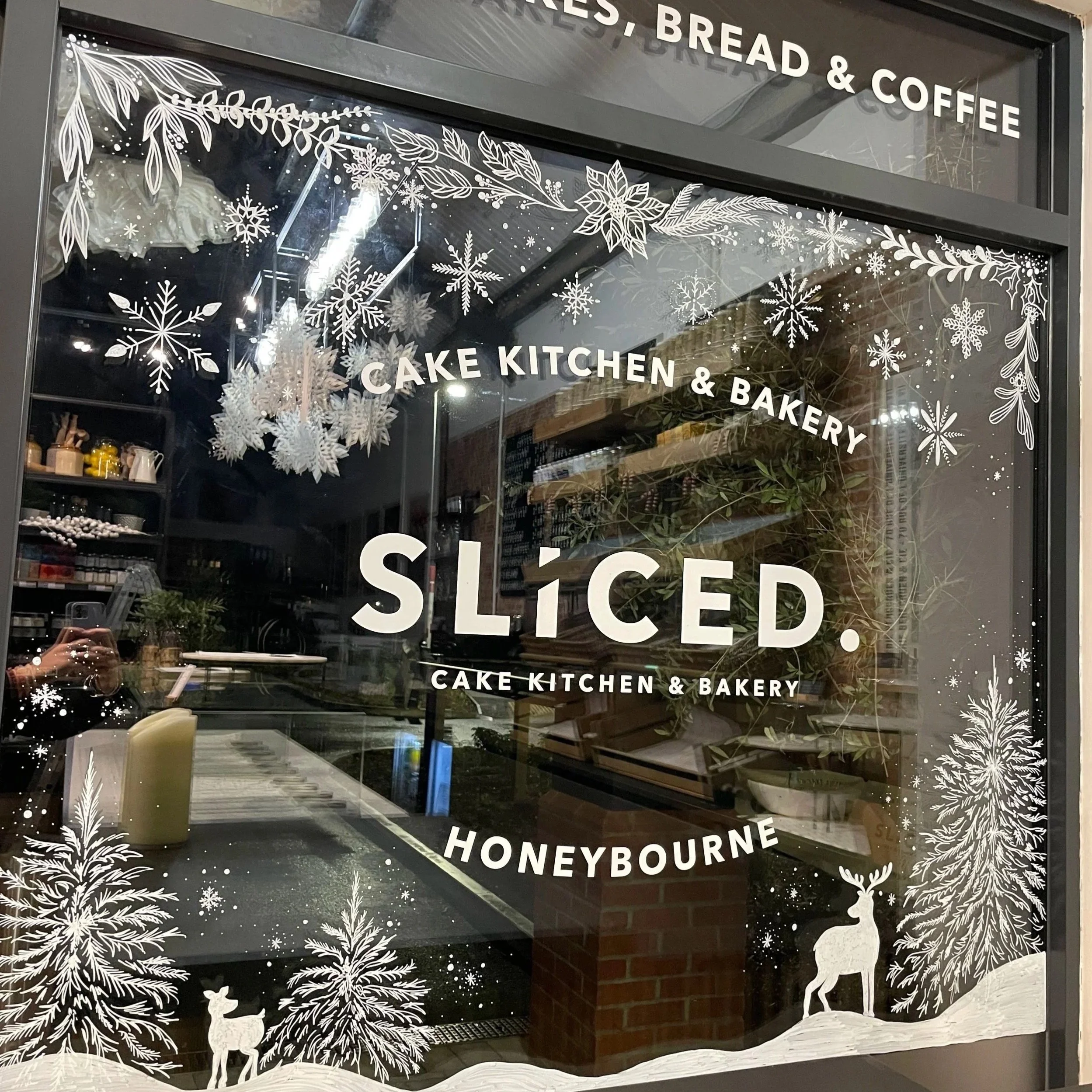 Decorative holiday-style window illustrations with white snowflakes, pine trees, and reindeer silhouettes, featuring text promoting a cake kitchen and bakery in a cafe setting.