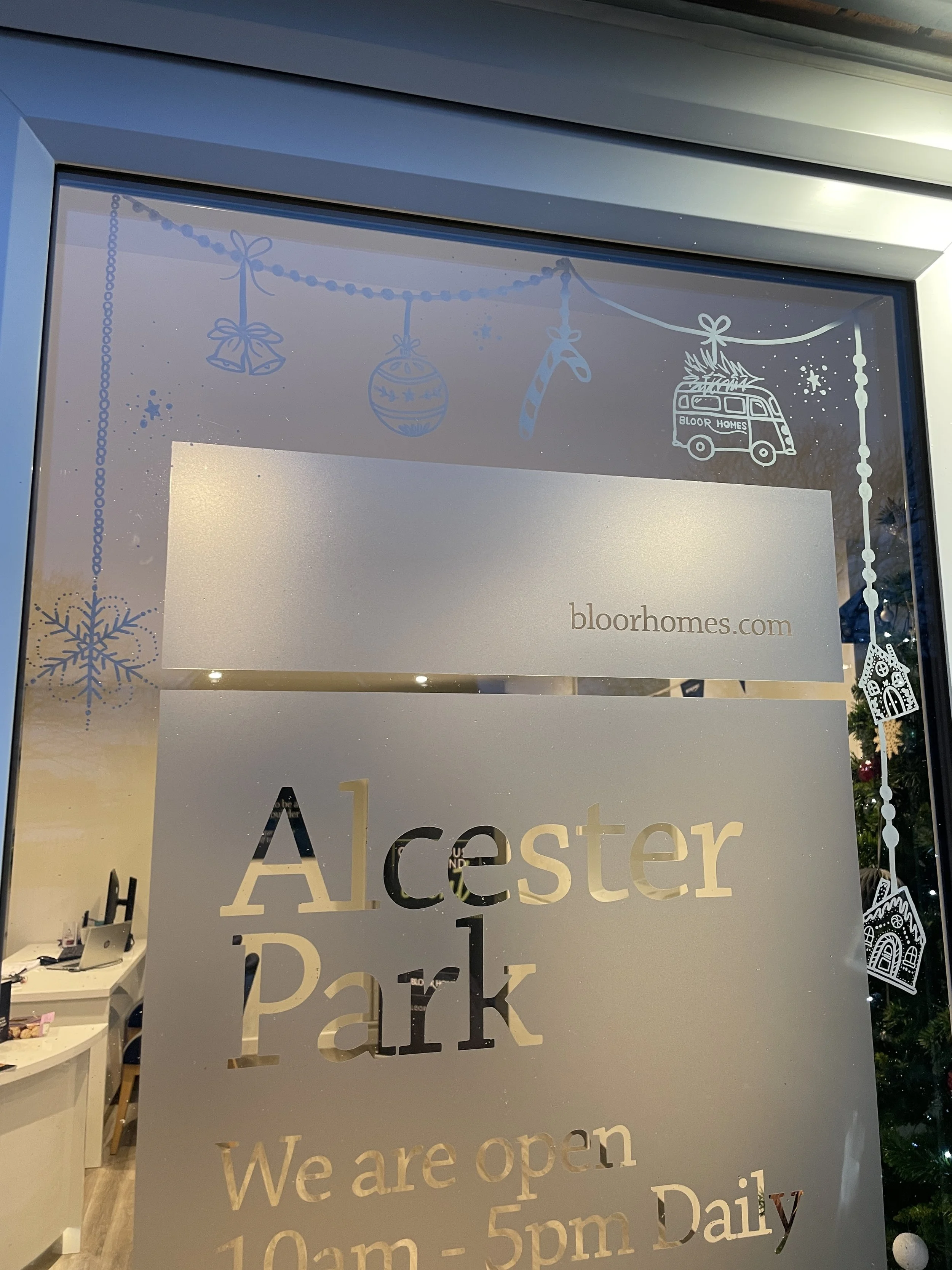 Glass door with a sign reading 'Alcester Park' and opening hours, decorated with Christmas-themed stickers including snowflakes, ornaments, a candy cane, a house, and a Christmas tree with presents.