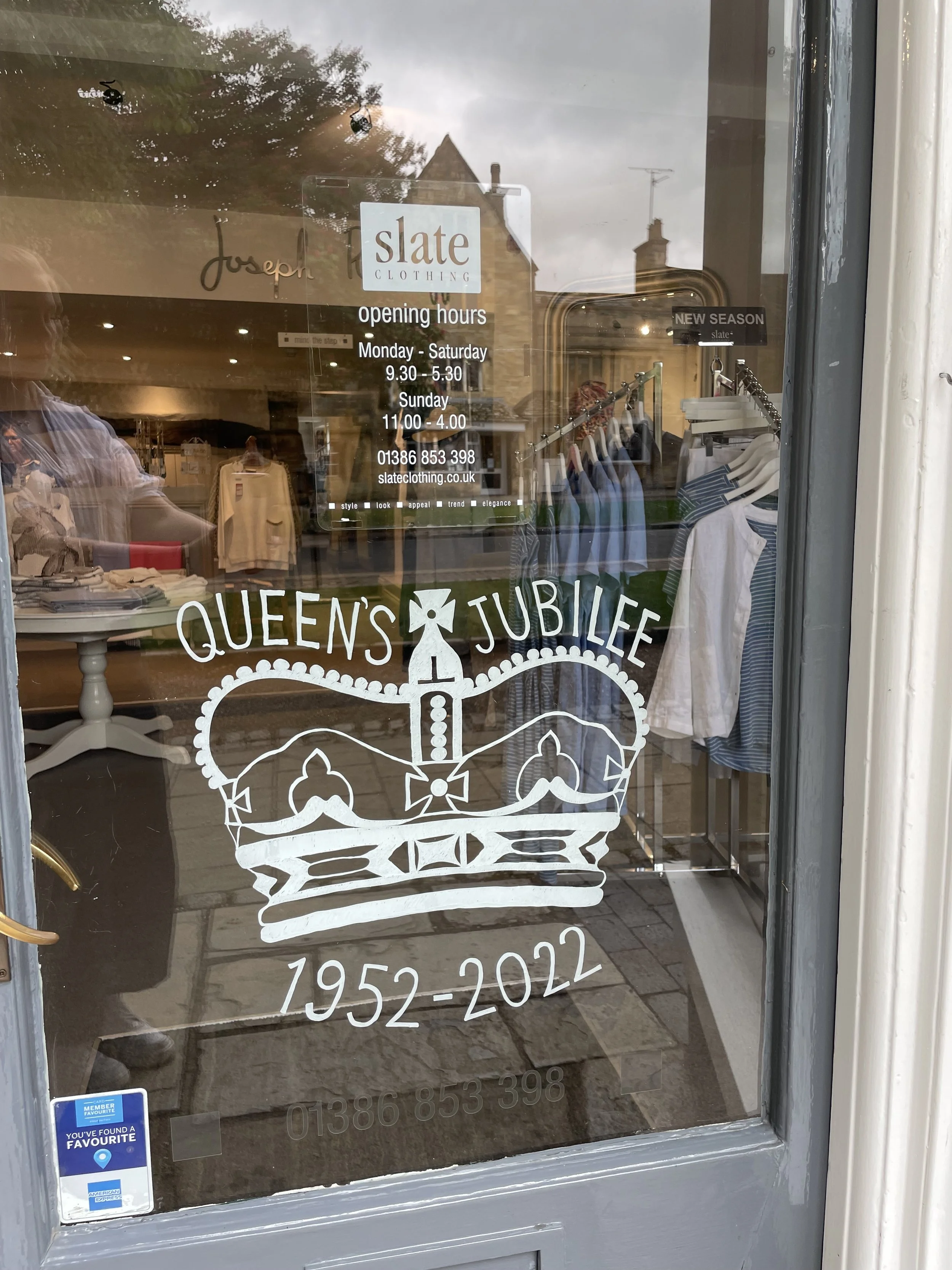 Storefront window with Queen's Jubilee celebration graphic of a crown above a ribbon and the text "Queen's Jubilee 1952-2022." Reflection of inside store and street outside visible.